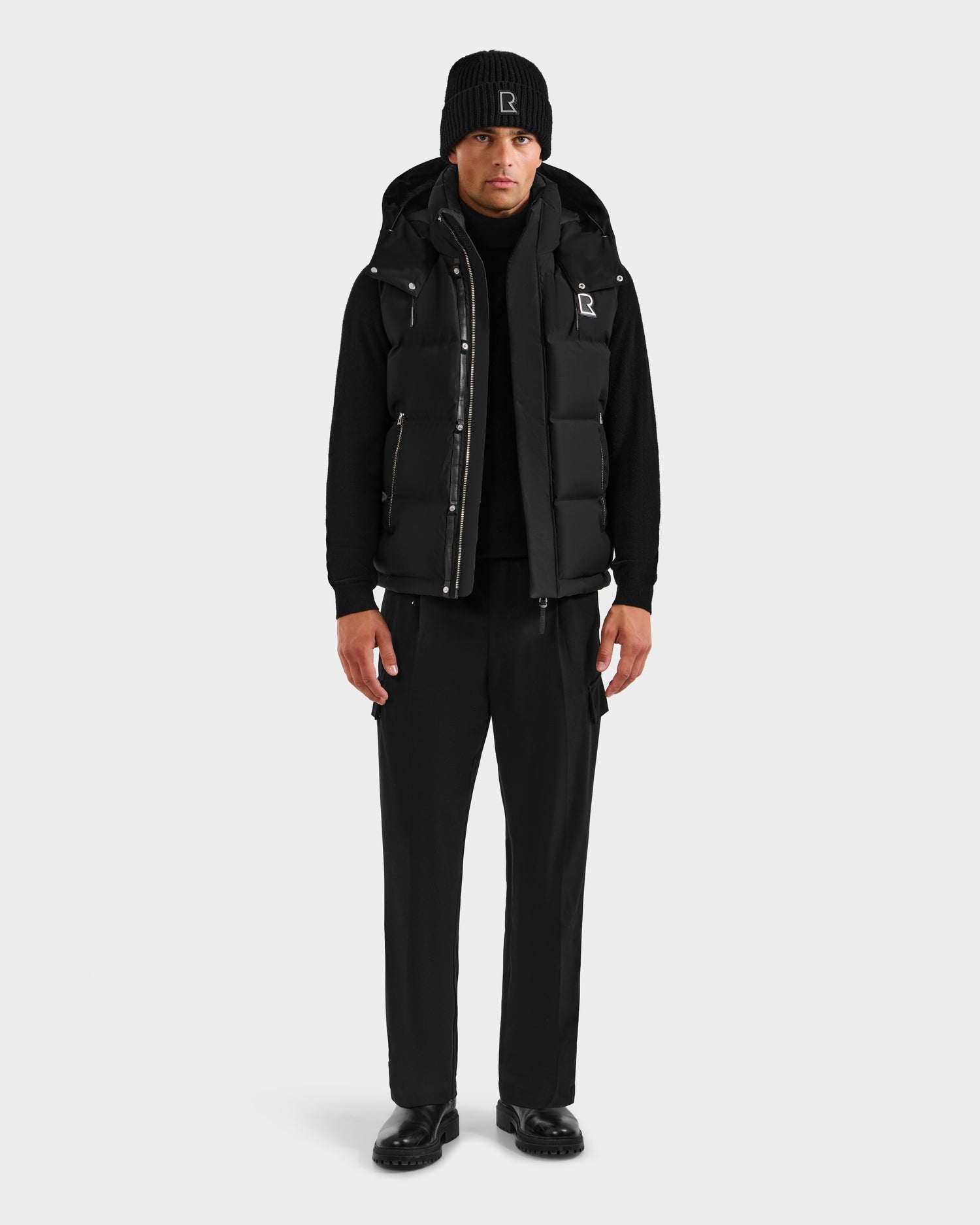A person faces forward, wearing the Rudsak REN H - 6125688 Black men’s quilted down puffer vest with a removable hood, styled with a black beanie, long-sleeve shirt, pants, and shoes against a plain light background.