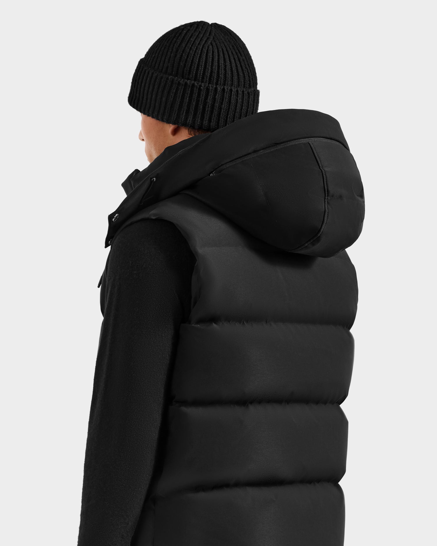 A person is seen from the back wearing the Rudsak REN H - 6125688 Black men's quilted down puffer vest with removable hood and a black knit beanie, set against a plain light gray background—a cold-weather essential.