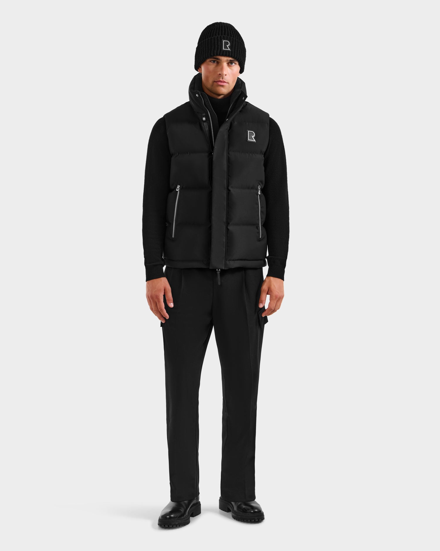 A man stands facing forward, wearing the Rudsak REN H - 6125688 Black men’s quilted down puffer vest with logo R—an essential cold-weather piece with a simple, modern design—paired with a black beanie, zip-up top, pants, and shoes.