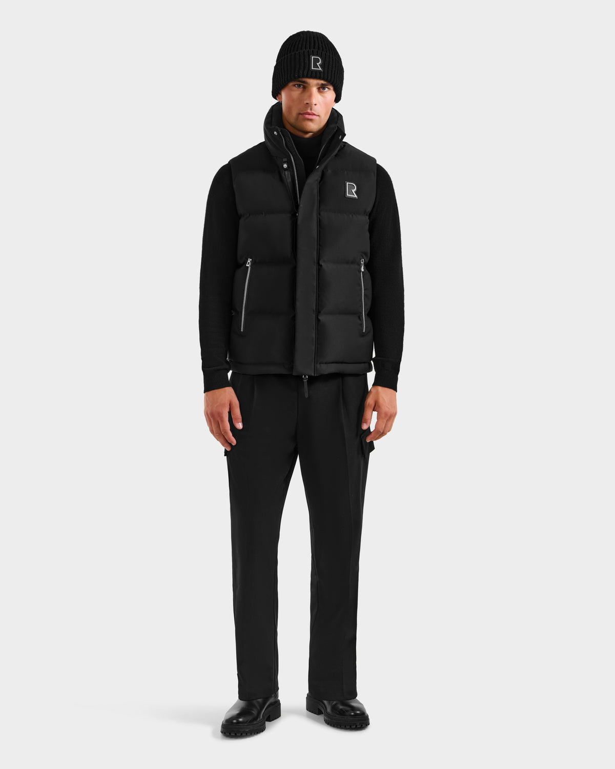 A man stands facing forward, wearing the Rudsak REN H - 6125688 Black men’s quilted down puffer vest with logo R—an essential cold-weather piece with a simple, modern design—paired with a black beanie, zip-up top, pants, and shoes.