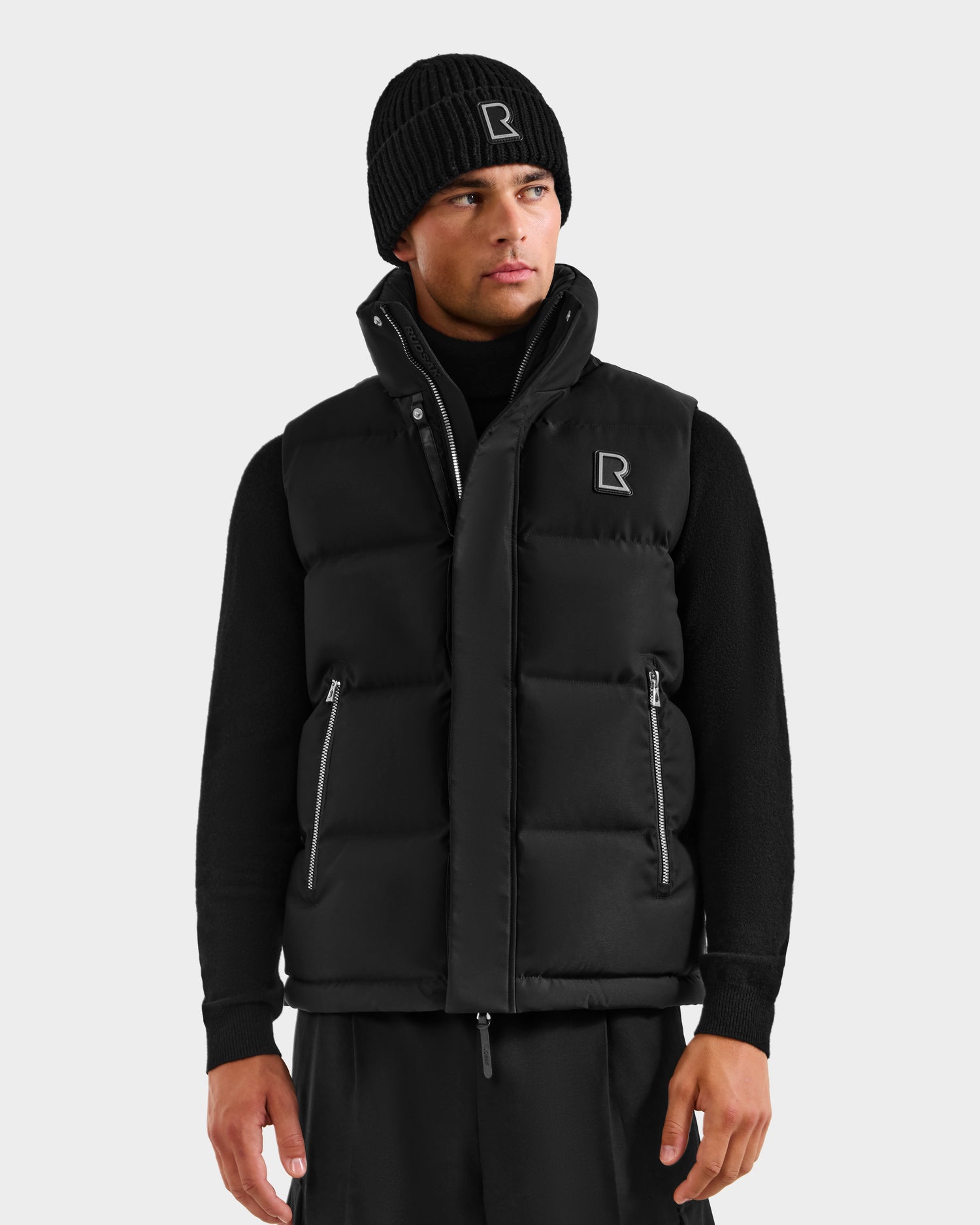 A man wears the Rudsak REN H - 6125688 Black beanie and men’s quilted down puffer vest, both featuring the white “R” logo, standing against a plain light gray background.