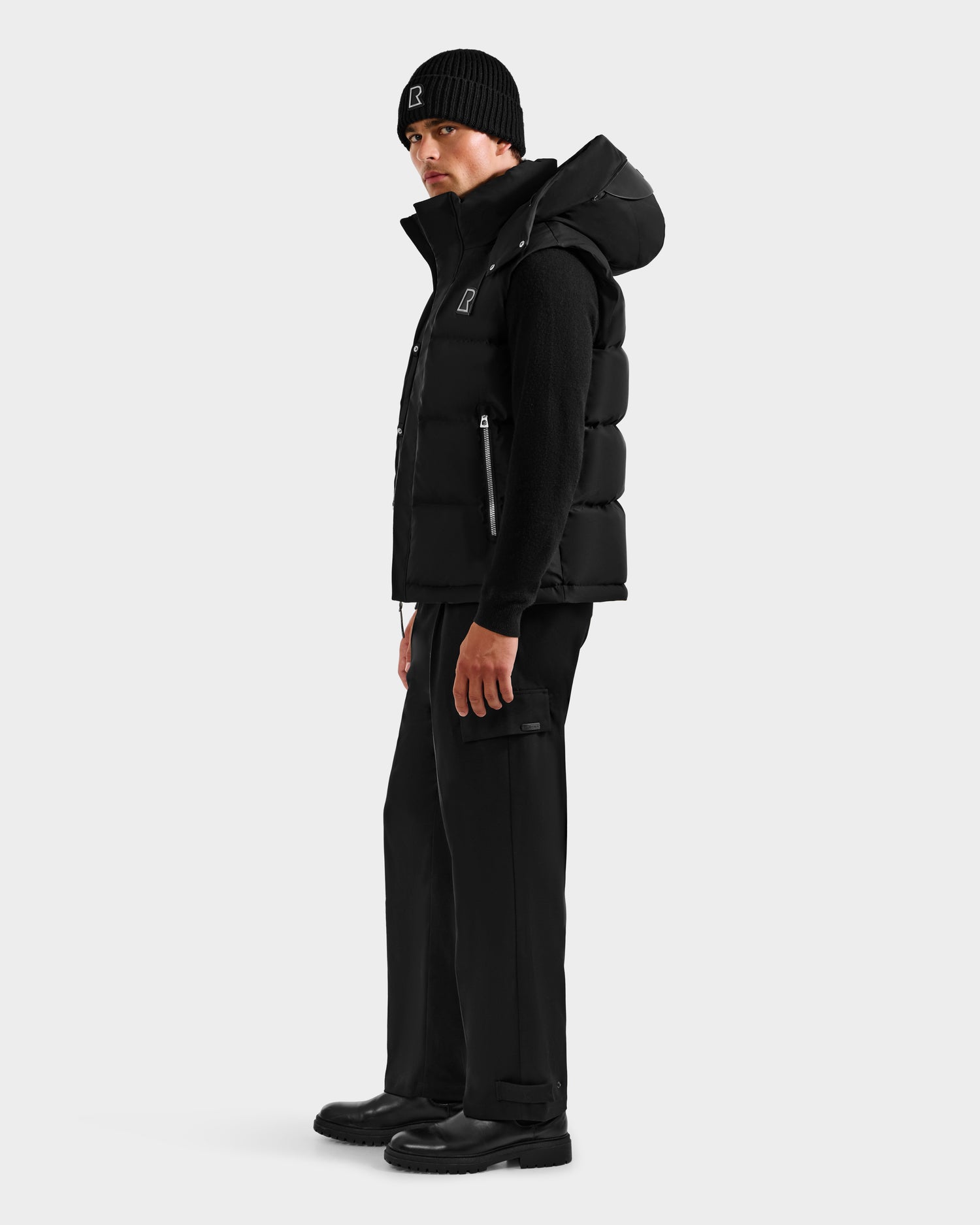 A man in a black Rudsak REN H - 6125688 beanie, quilted down puffer vest with removable hood, long-sleeve top, cargo pants, and boots stands in profile against a light background—a true cold-weather essential look.