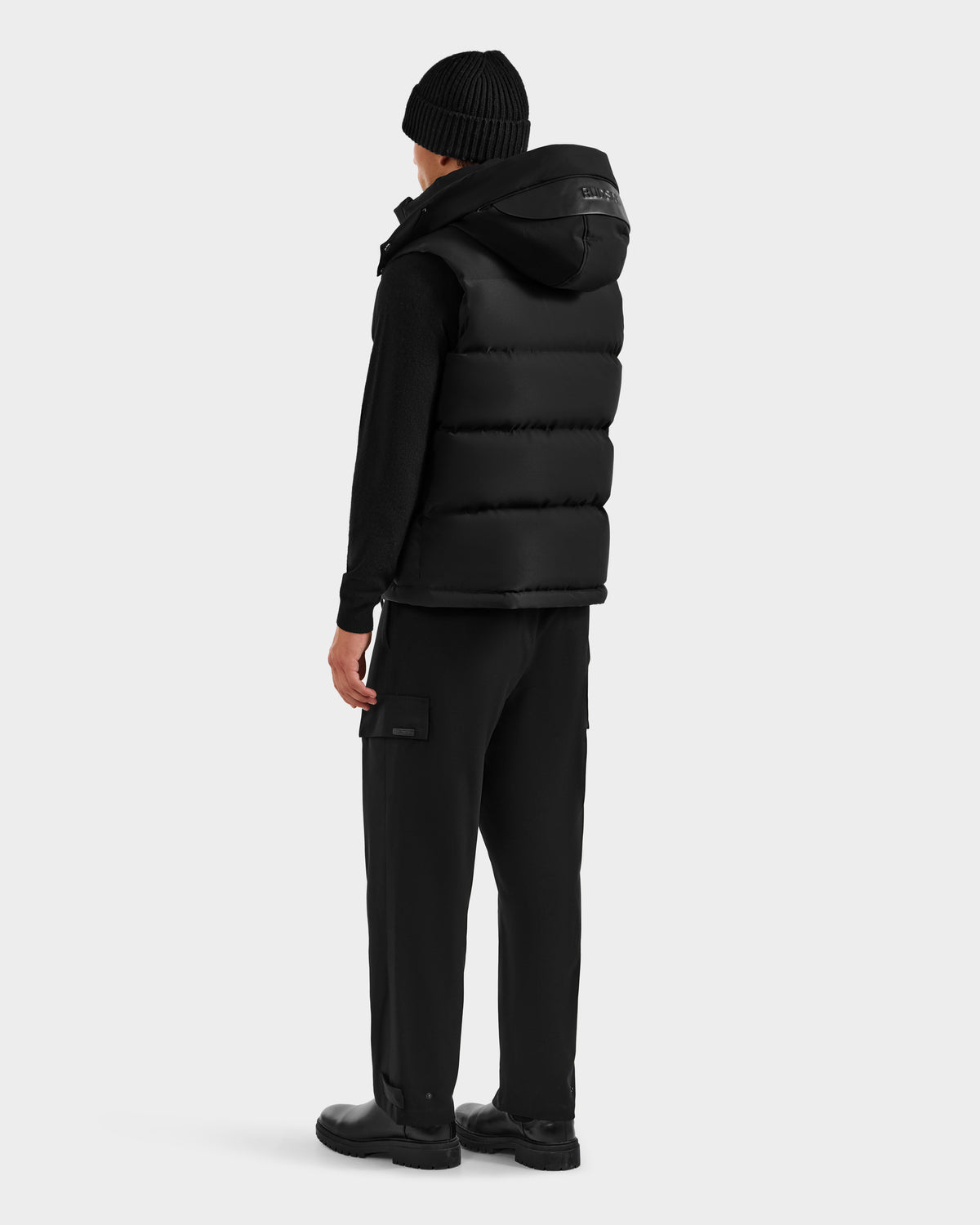 A person faces away, wearing the REN H - 6125688 Black quilted down puffer vest with removable hood by Rudsak over a black long-sleeve top, cargo pants, and shoes, set against a plain light gray background—a cold-weather essential.