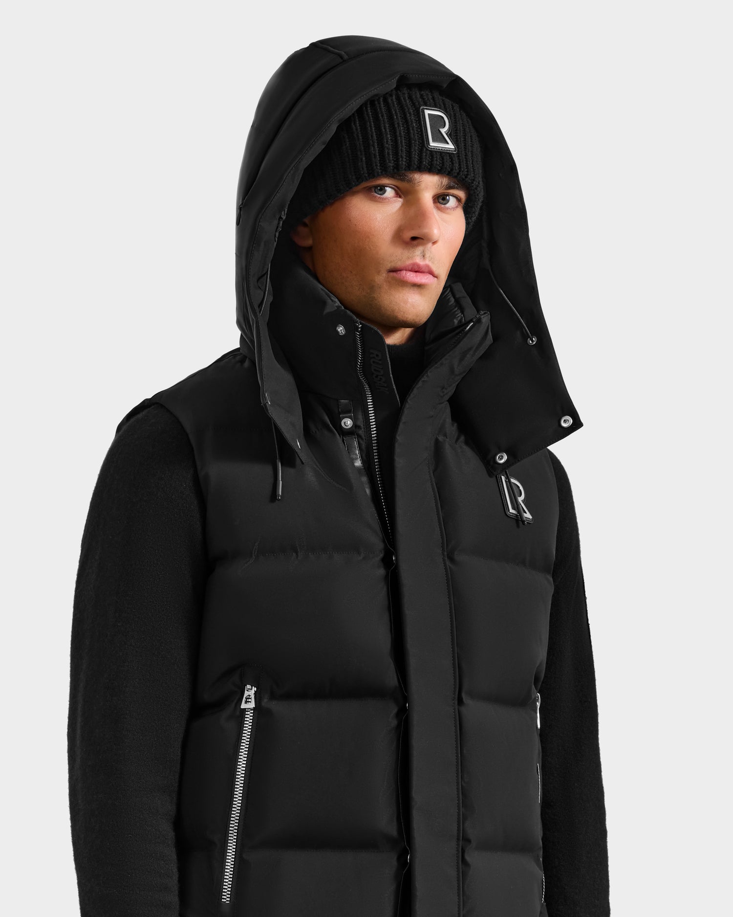 A young man models the Rudsak REN H - 6125688 Black men’s quilted down puffer vest with a removable hood, layered over a black sweater and a black beanie with a white R logo—perfect for cold weather. He stands against a light gray background.