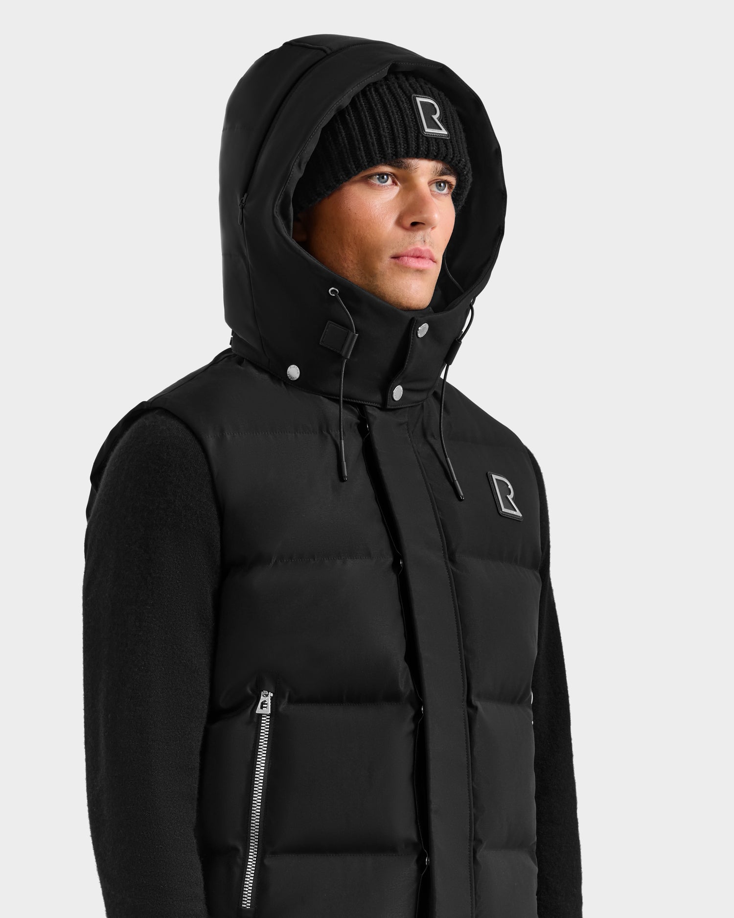 A man stands against a plain light background wearing the black Rudsak REN H - 6125688 men’s quilted down puffer vest with zippered pocket and removable hood, paired with a matching black knit beanie—both featuring the white R logo.