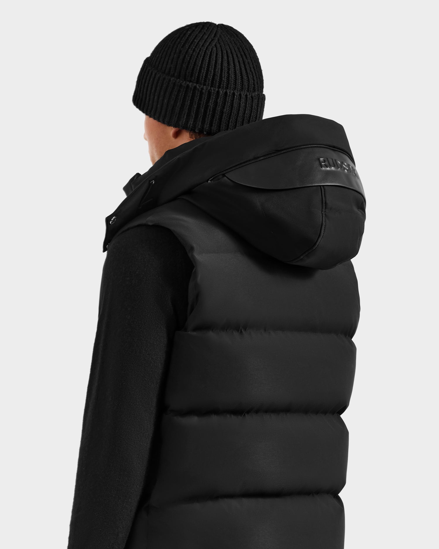 A person wearing the Rudsak REN H - 6125688 Black men’s quilted down puffer vest with a removable hood and a black beanie, viewed from behind against a plain light background—an ideal cold-weather essential.