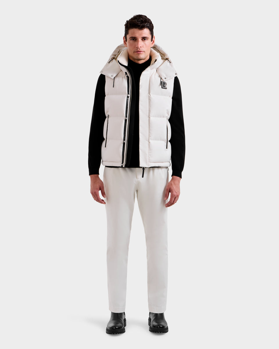 A man faces forward wearing the Rudsak REN H - 6125688 Vanilla quilted down vest over a black long-sleeve shirt, white drawstring pants, and black shoes, set against a plain light background, highlighting this cold-weather essential.