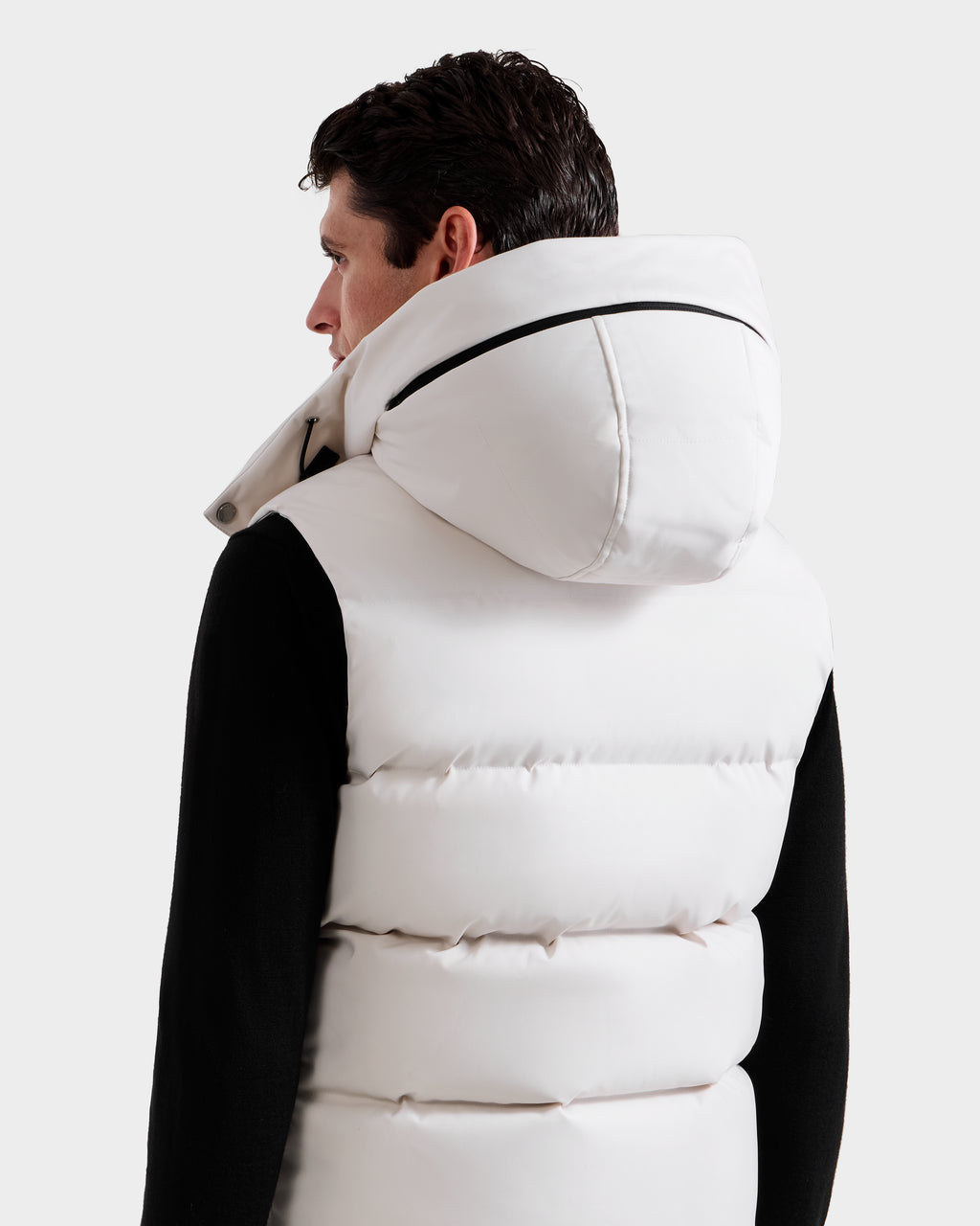 A person with short dark hair is seen from behind, wearing the Rudsak REN H - 6125688 Vanilla men’s quilted down vest with the hood up over a black long-sleeve shirt, set against a plain light gray background.