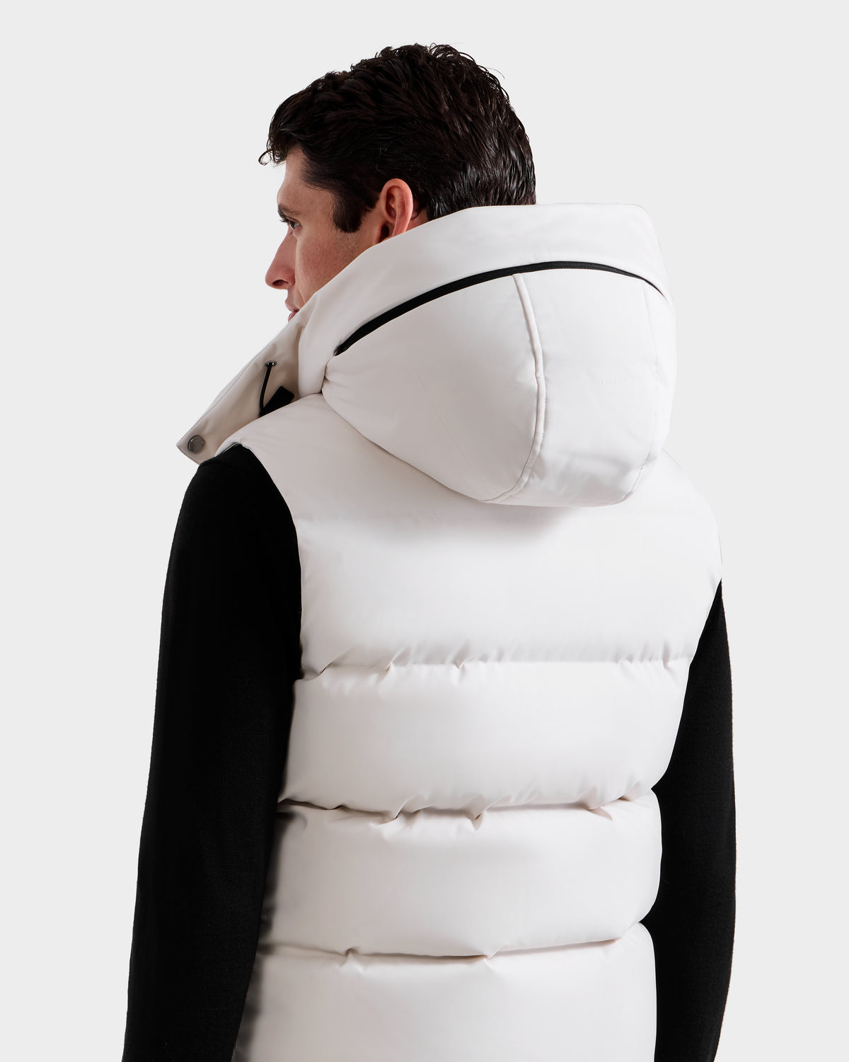 A person with short dark hair is seen from behind, wearing the Rudsak REN H - 6125688 Vanilla men’s quilted down vest with the hood up over a black long-sleeve shirt, set against a plain light gray background.
