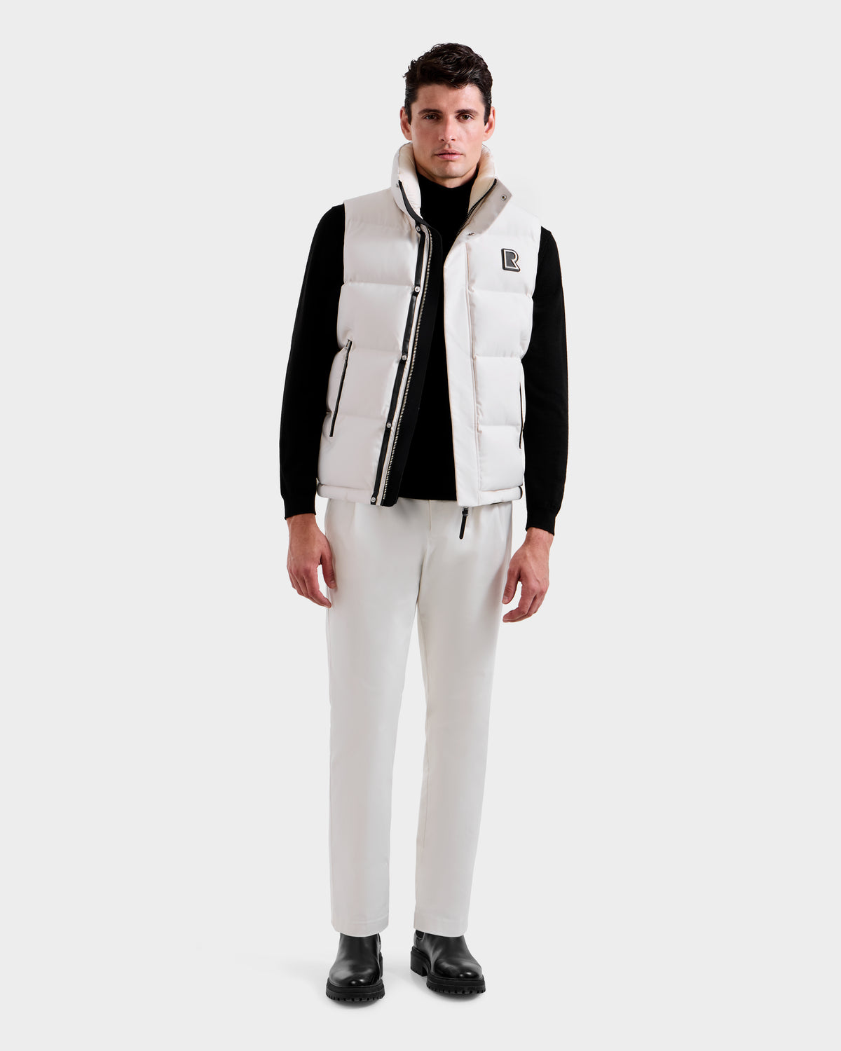 A man faces forward in a Rudsak REN H - 6125688 Vanilla quilted down vest over a black long-sleeve shirt, white pants, and black boots, highlighting this cold-weather essential against a plain light background.