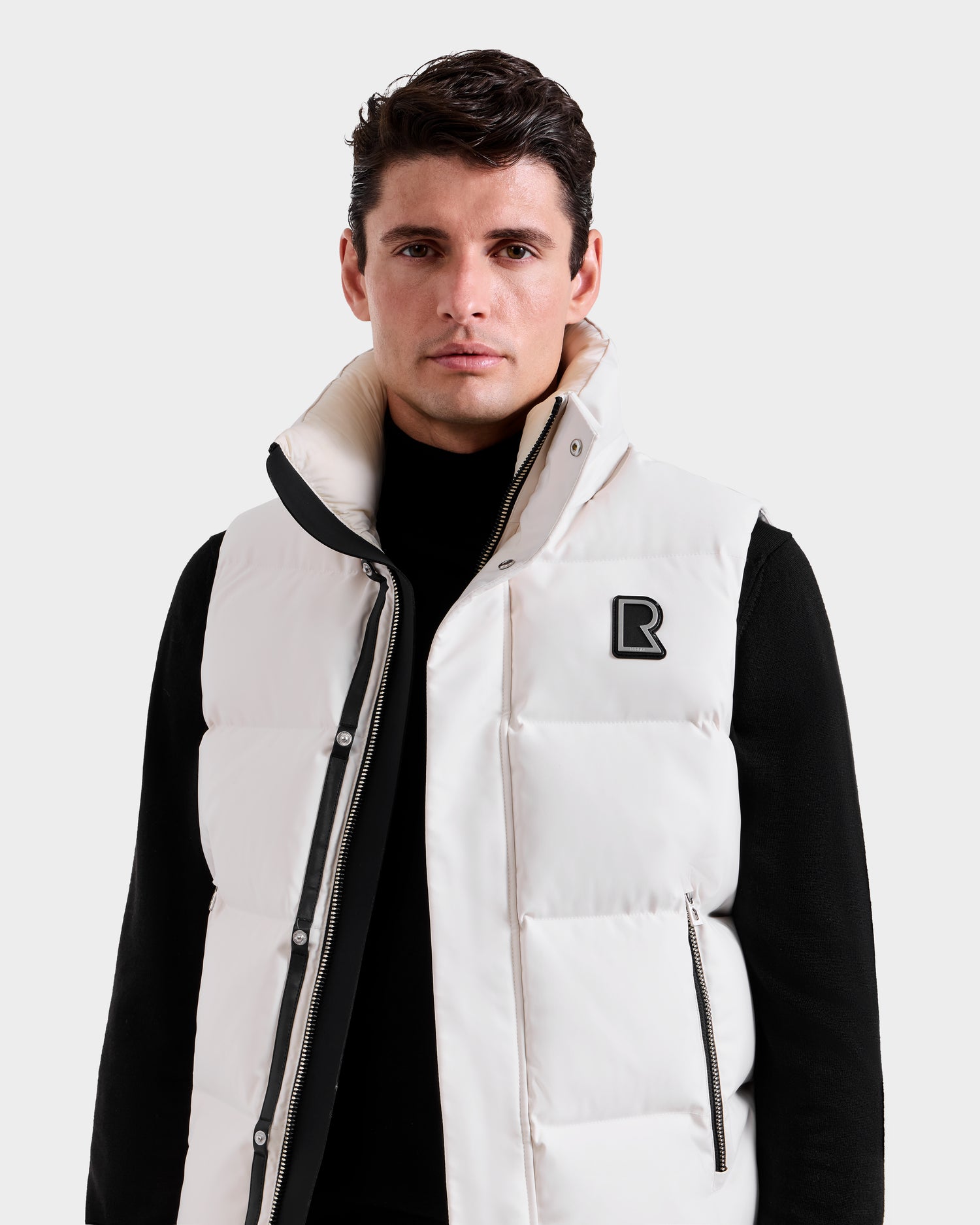 A man with short dark hair wears the Rudsak REN H - 6125688 Vanilla men’s quilted down vest over a black turtleneck, standing against a plain light gray background—a stylish cold-weather essential.
