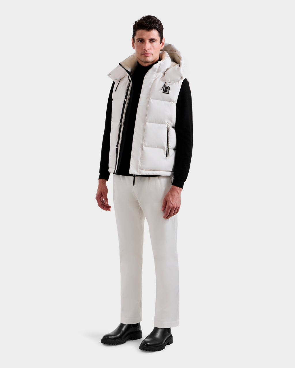 A man faces forward against a plain light background, wearing the Rudsak REN H - 6125688 Vanilla men’s quilted down vest with hood—styled with a black long-sleeve shirt, white pants, and black boots for cold-weather appeal.