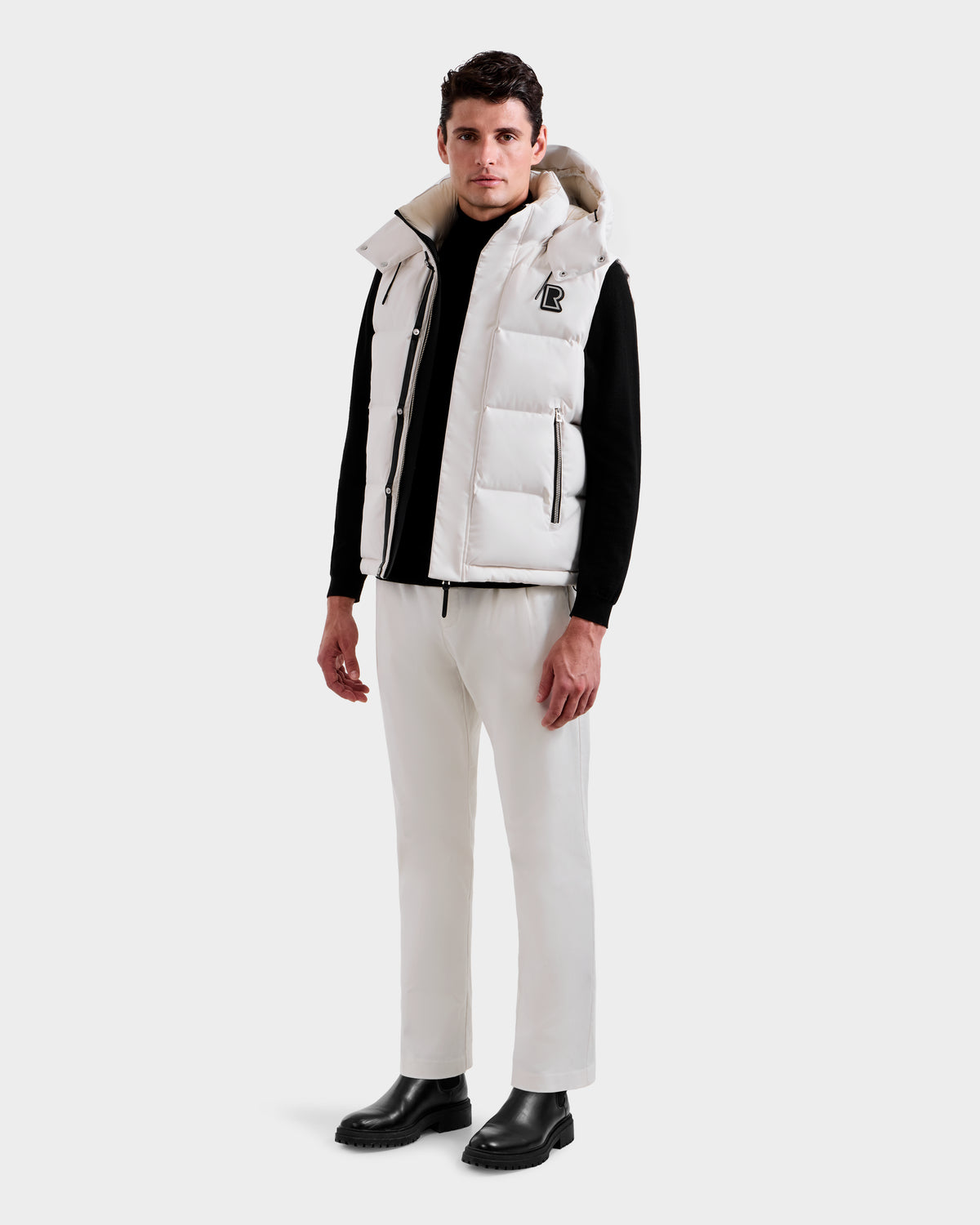 A man faces forward against a plain light background, wearing the Rudsak REN H - 6125688 Vanilla men’s quilted down vest with hood—styled with a black long-sleeve shirt, white pants, and black boots for cold-weather appeal.