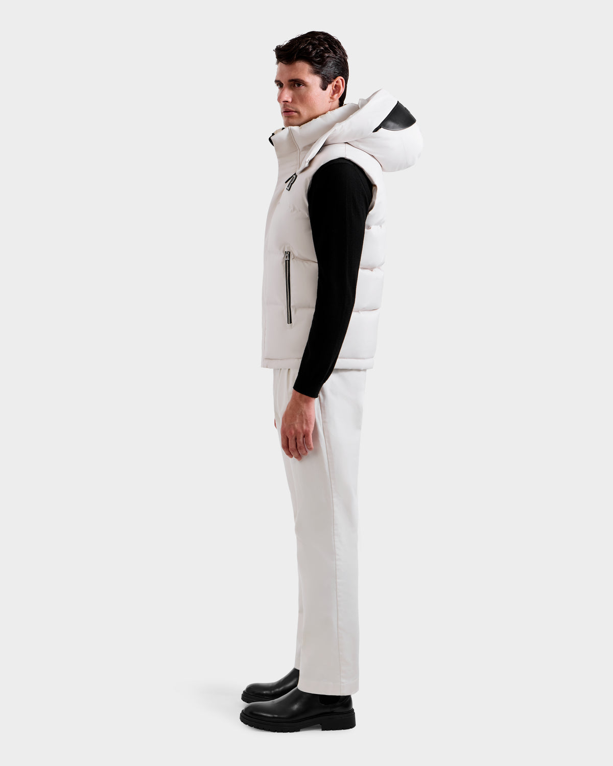 A man in profile wears the Rudsak REN H - 6125688 Vanilla quilted down vest with a hood, styled with a black long-sleeve shirt, white pants, and black boots, standing against a plain light gray background.