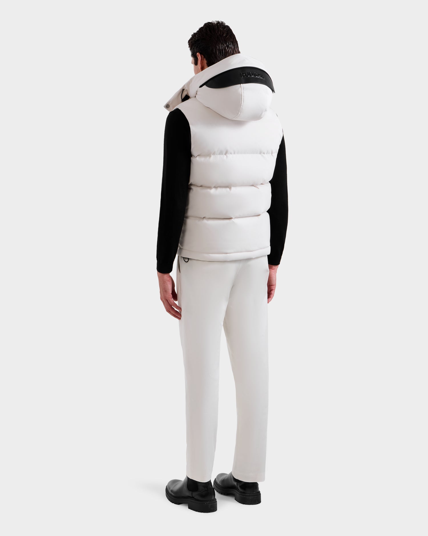 A man faces away wearing the Rudsak REN H - 6125688 Vanilla men’s quilted down puffer vest with hood, paired with a black shirt and white pants against a white background—a true cold-weather essential.