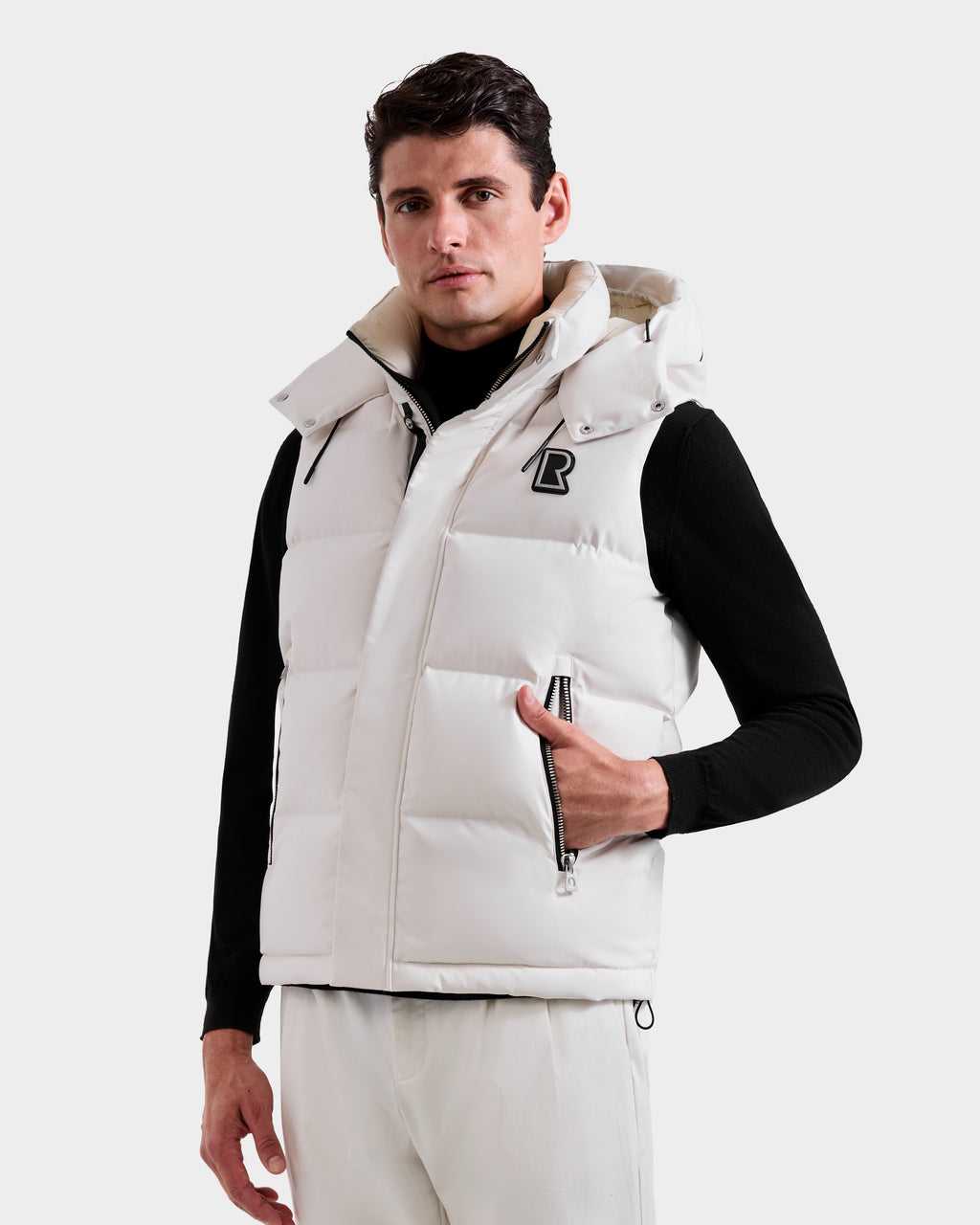 A man faces forward in a Rudsak REN H - 6125688 Vanilla men's quilted down puffer vest with removable hood, zippered pockets, and B logo on the chest, layered over a black turtleneck and white pants against a plain white background.