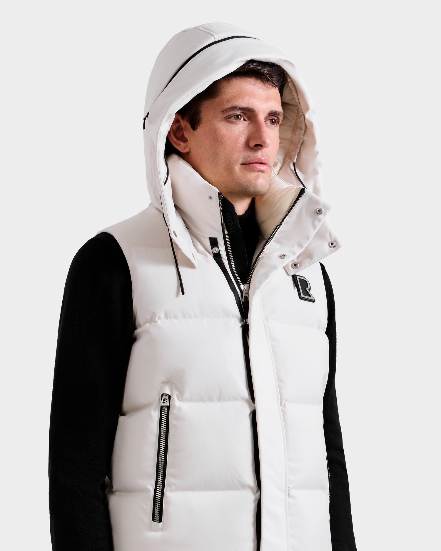 A man in a Rudsak REN H - 6125688 Vanilla men’s quilted down vest with hood and black zipper over a black long-sleeve shirt, looking right against a plain background—an ideal cold-weather essential.
