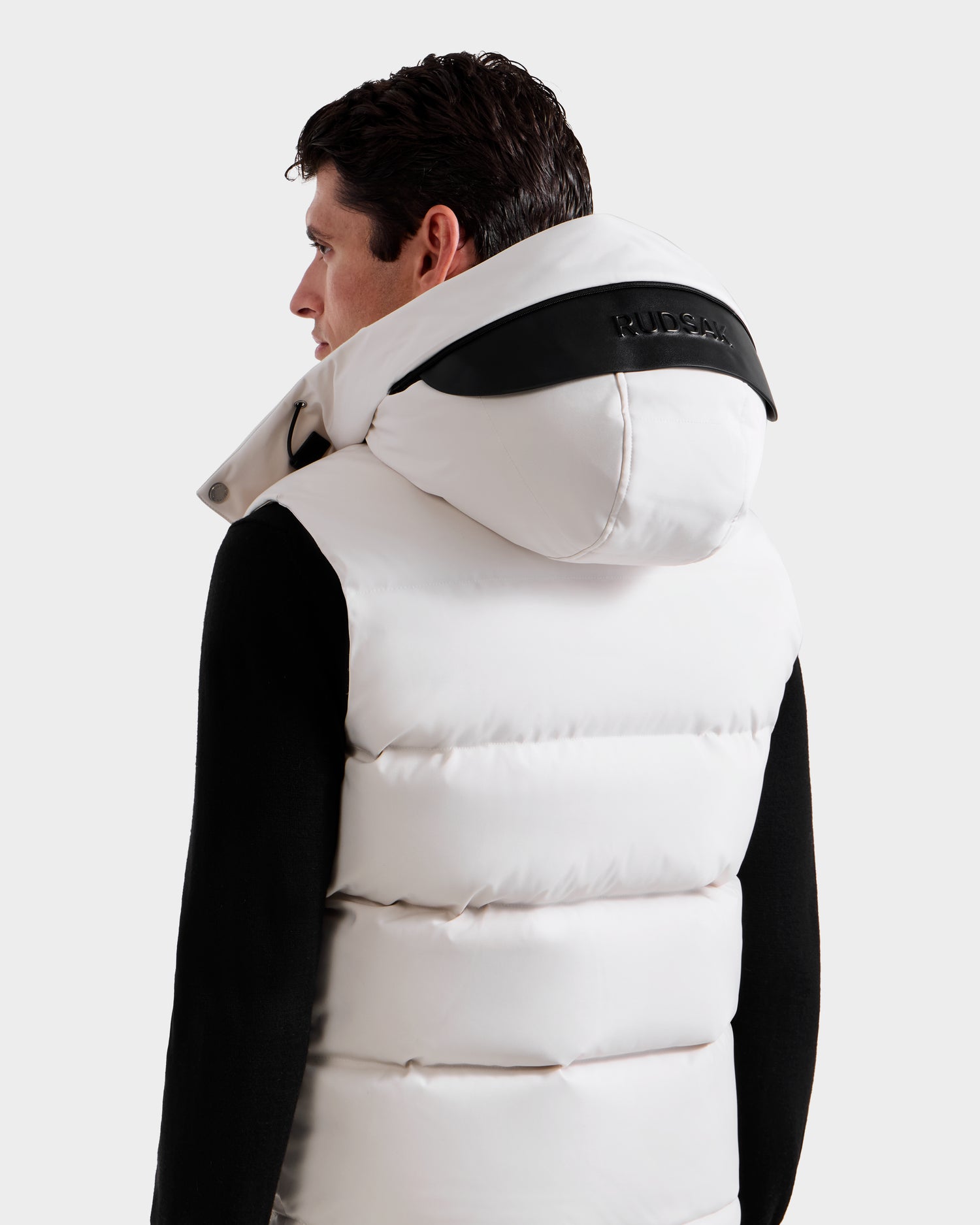 A man stands facing away, wearing the Rudsak REN H - 6125688 Vanilla men’s quilted down vest with a large hood and black detailing, layered over a black long-sleeve shirt, showcasing the brand’s cold-weather style.