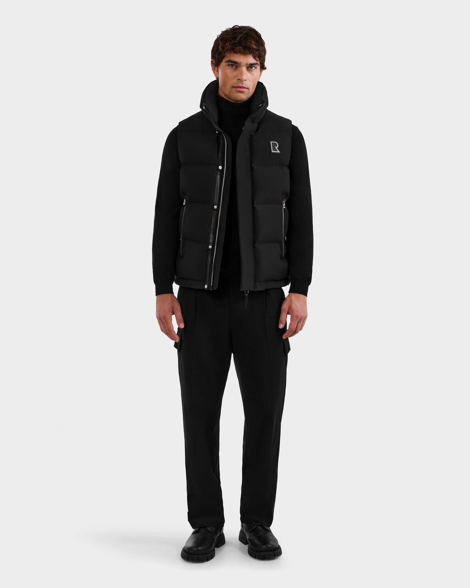 A man faces forward wearing the Rudsak REN M - 6125691 Black men’s quilted down puffer vest over a black turtleneck, pants, and shoes, set against a plain light background.