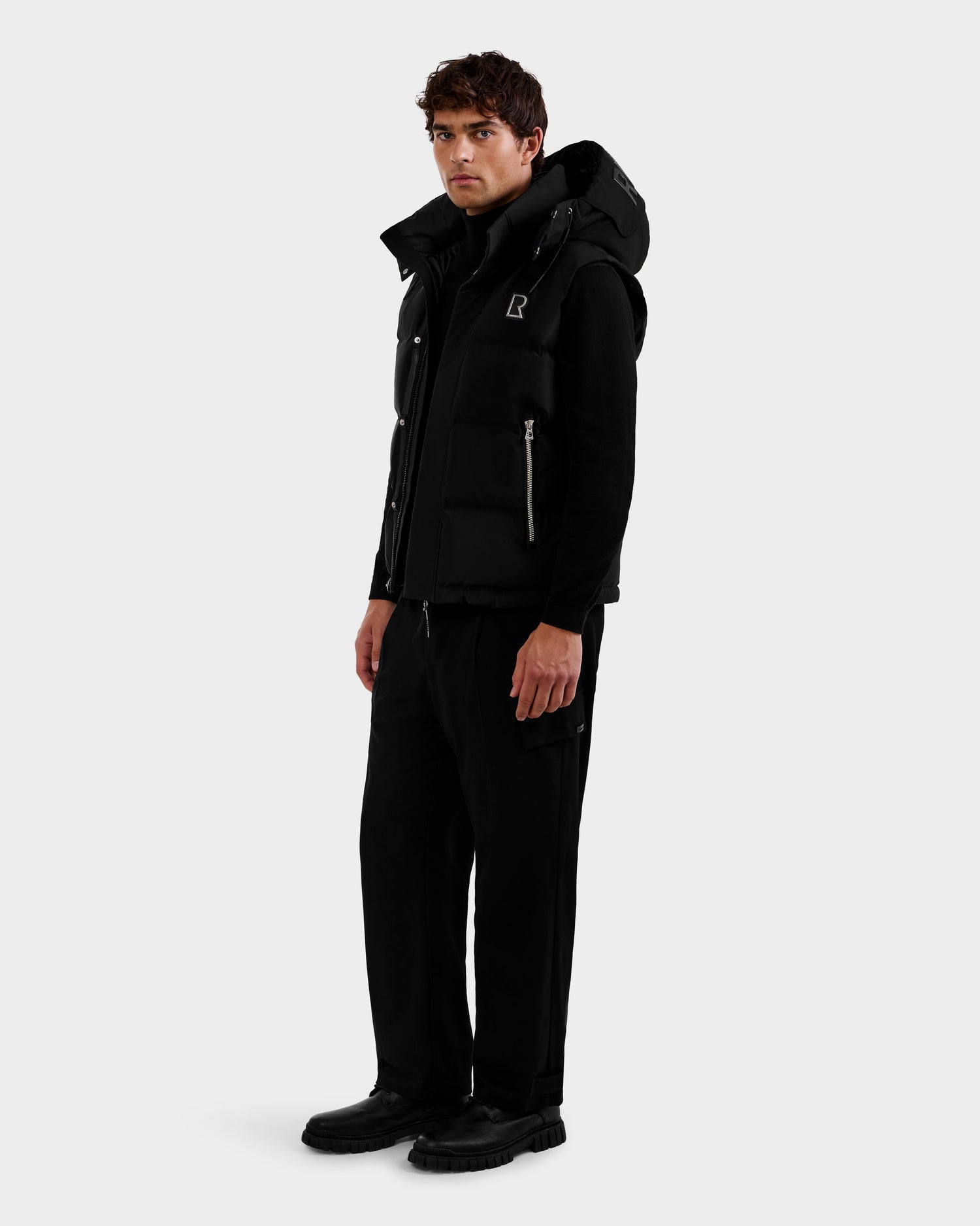 A man stands slightly to the side, wearing the Rudsak REN M - 6125691 Black men's quilted down puffer vest over a black outfit with cargo pants and black shoes, against a plain light gray background.