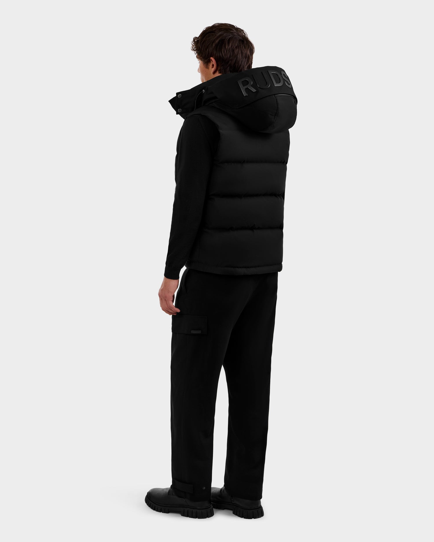 A person faces away, wearing the Rudsak REN M - 6125691 Black men's quilted down puffer vest with bold RUD lettering on the hood, styled with black long sleeves, cargo pants, and shoes against a plain light background.