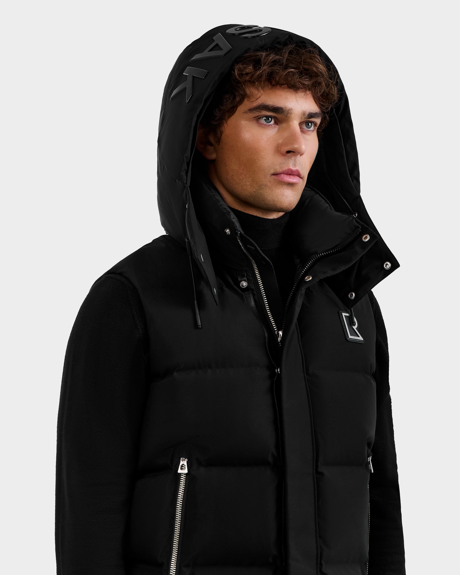 A man wears the Rudsak REN M - 6125691 Black men's quilted down puffer vest, unzipped over a black long-sleeve top. This cold-weather essential features a hood with bold printed letters, set against a light gray background.