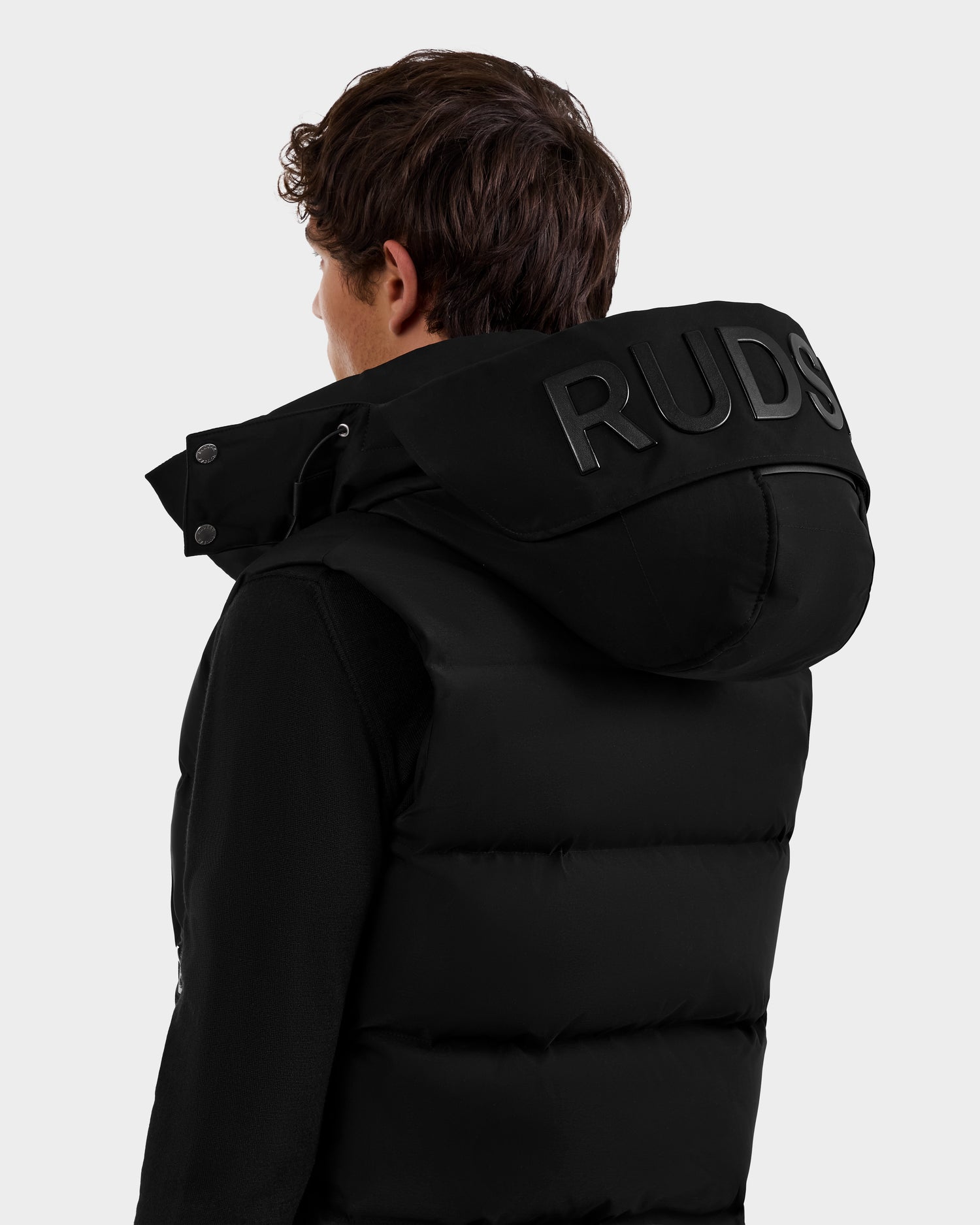 A person with short brown hair is seen from the back wearing a black Rudsak REN M - 6125691 men’s quilted down puffer vest with a large hood featuring bold, raised “RUDS” letters. The background is plain light grey.