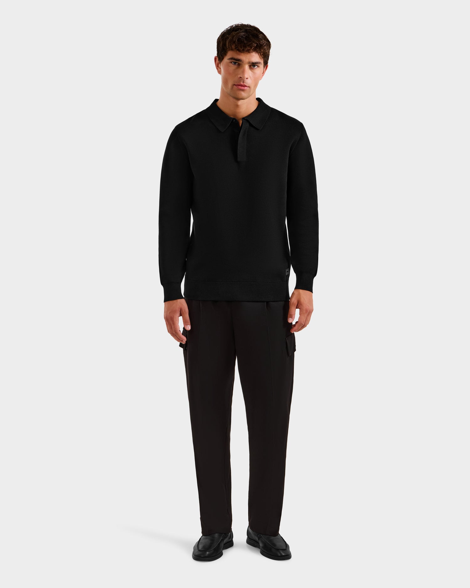 A man stands facing forward, wearing the Rudsak RICHARD - 6524529 black men's knit polo sweater with black trousers and shoes against a plain light gray background.