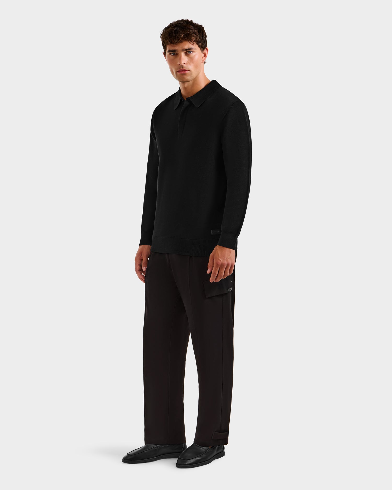 A man faces forward with a neutral expression, wearing the RUDSAK RICHARD - 6524529 Black men's knit polo sweater, loose dark brown side-pocket trousers, and black shoes, set against a plain background.