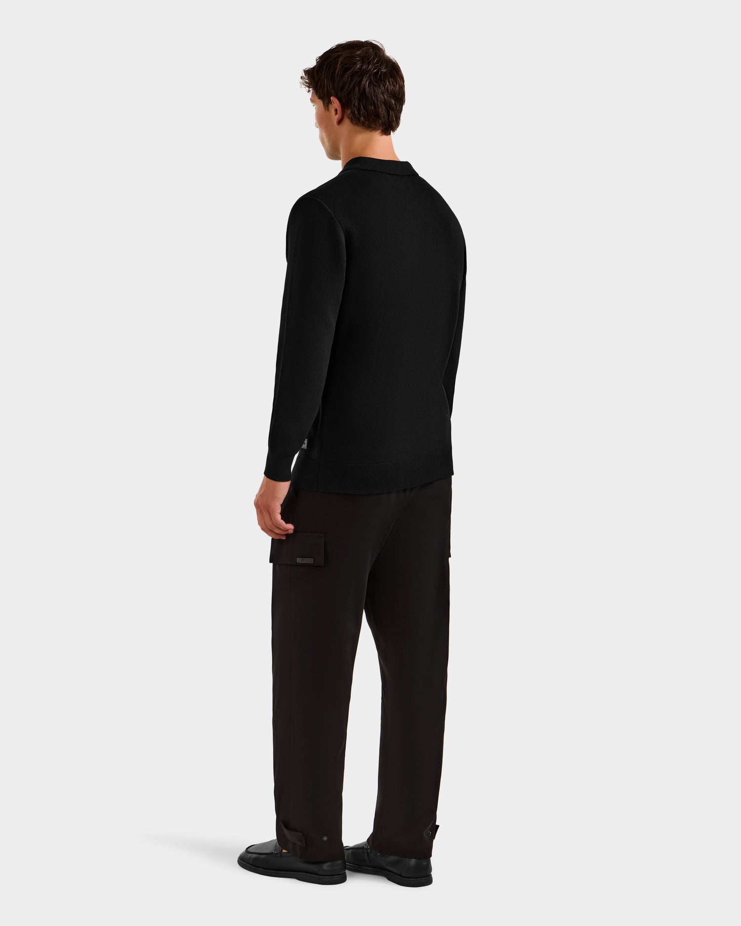 A man stands facing away, wearing the Rudsak RICHARD - 6524529 Black men's knit polo sweater, loose black cargo pants, and black shoes—an effortlessly modern staple against a plain white background.