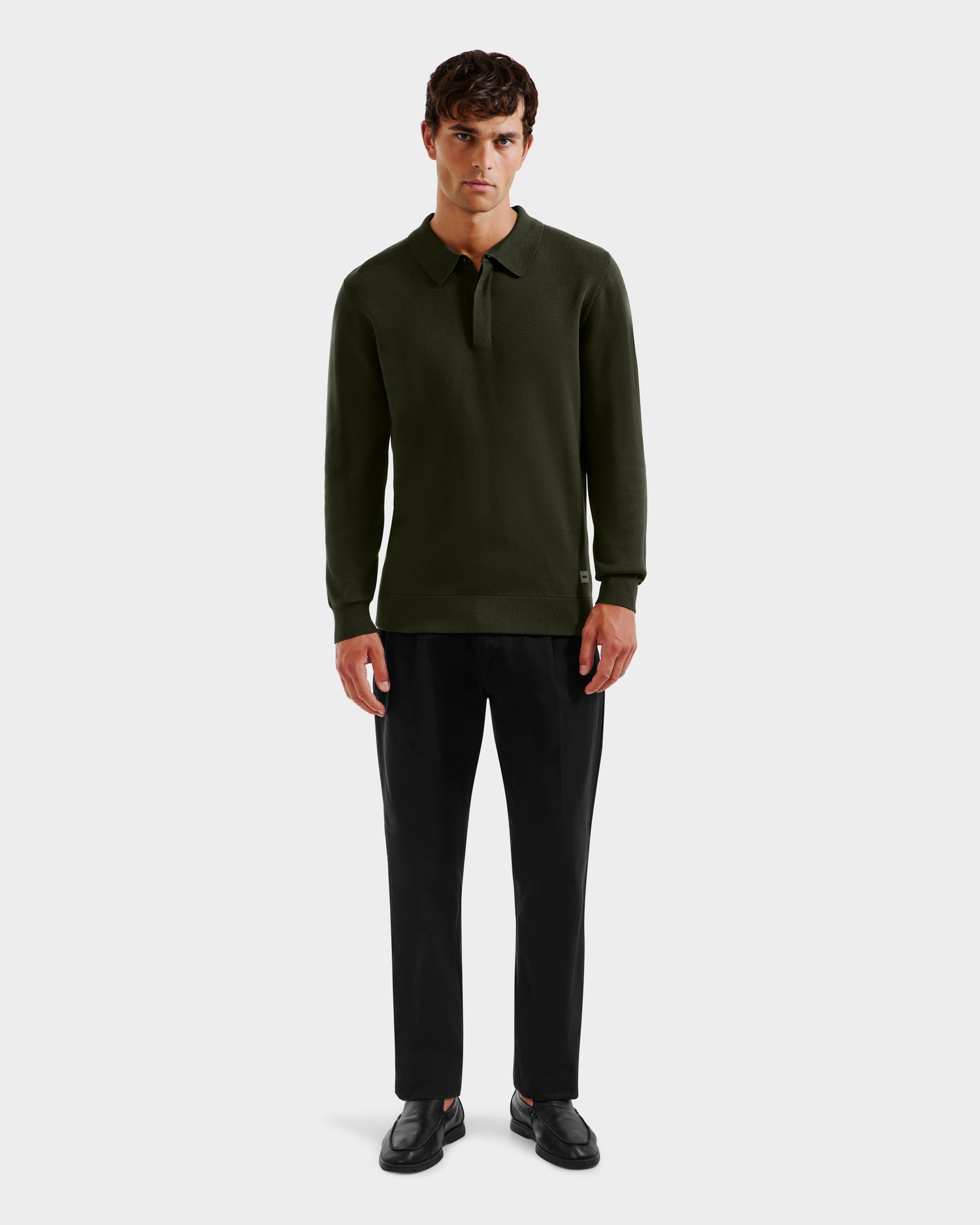 A man faces forward in a Rudsak RICHARD - 6524529 Dark Olive mens knit polo sweater, paired with black trousers and loafers, against a plain light gray background.