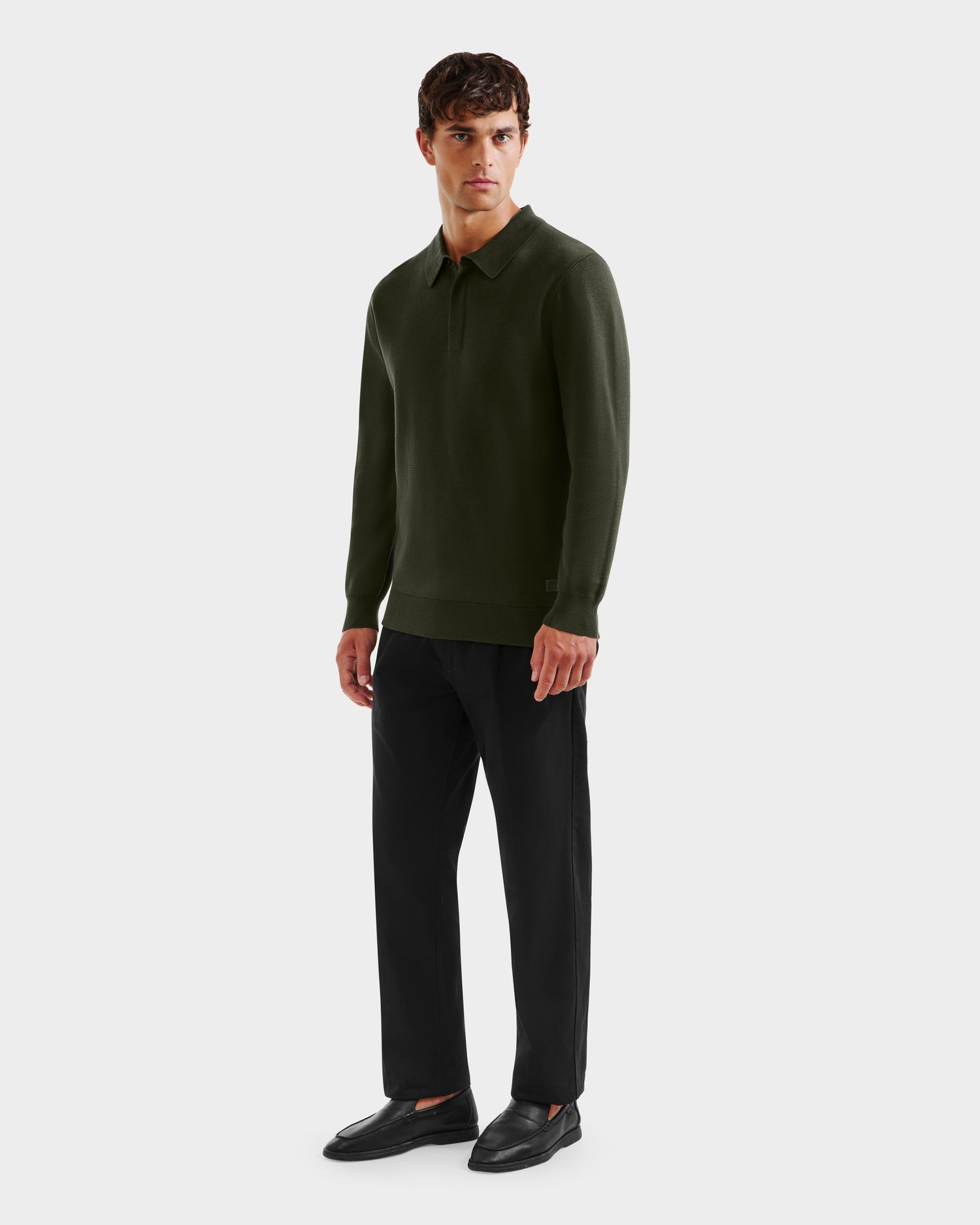 A man stands facing forward in a Rudsak RICHARD - 6524529 Dark Olive mens knit polo sweater, black pants, and black loafers against a plain light gray background.