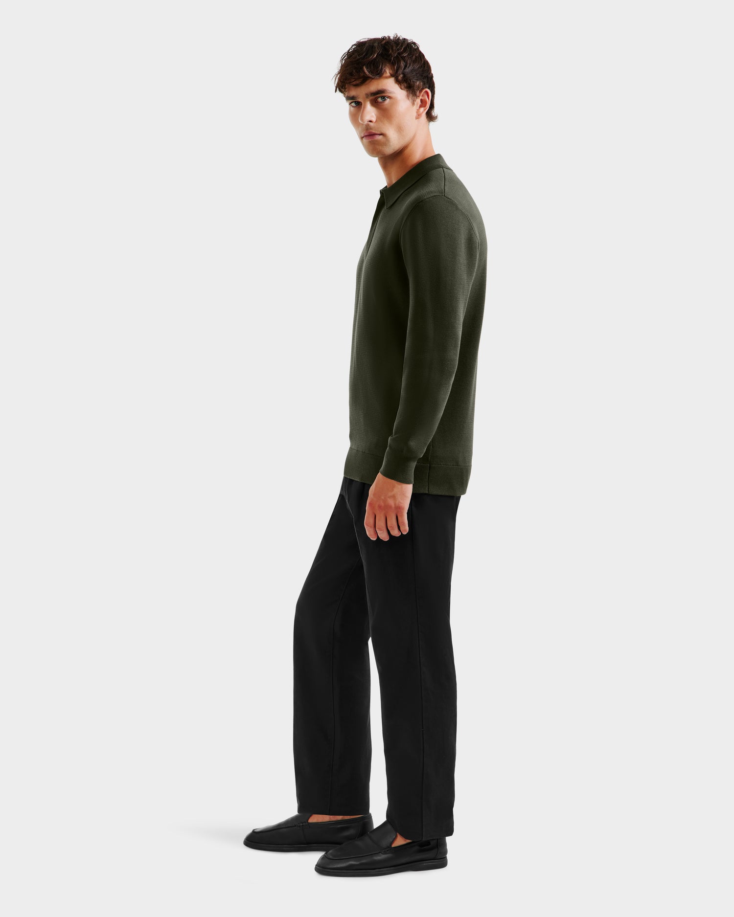 A man stands sideways against a plain white background, wearing the Rudsak RICHARD - 6524529 knit polo sweater in dark olive, paired with black pants and shoes. He gazes slightly toward the camera with a neutral expression.