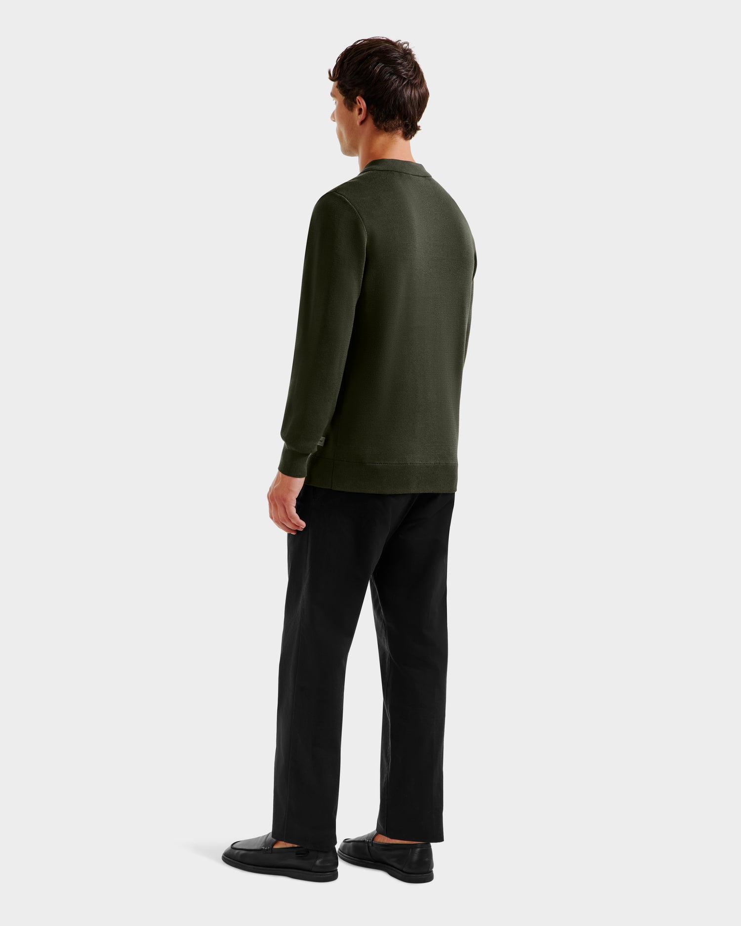 A person with short dark hair faces away from the camera, wearing the Rudsak RICHARD - 6524529 Dark Olive mens knit polo sweater, black pants, and black shoes against a plain light gray background.