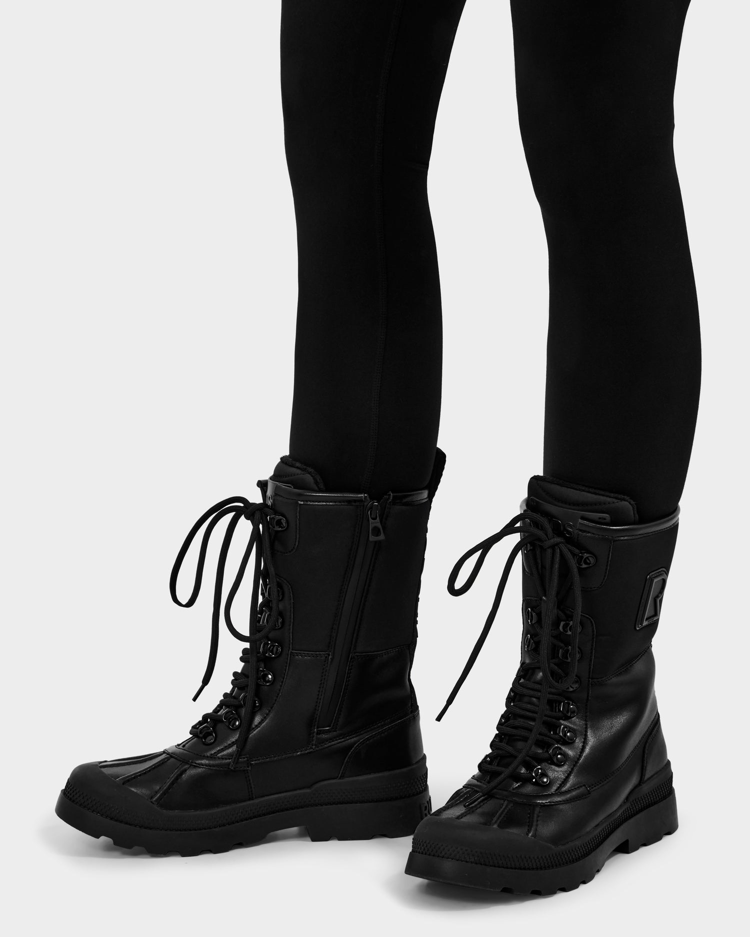 A person wearing Rudsak’s RITA - L222502 Black combat boots with side zippers and thick soles, paired with black leggings, stands on a light background.