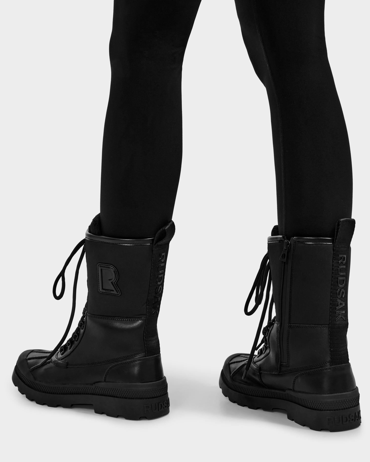 A person in black Rudsak RITA - L222502 women's lace-up winter boots with thick soles and weather-treated leather, wearing black leggings, shown from the back on a white background. The boots have the Rudsak logo on the side and back.