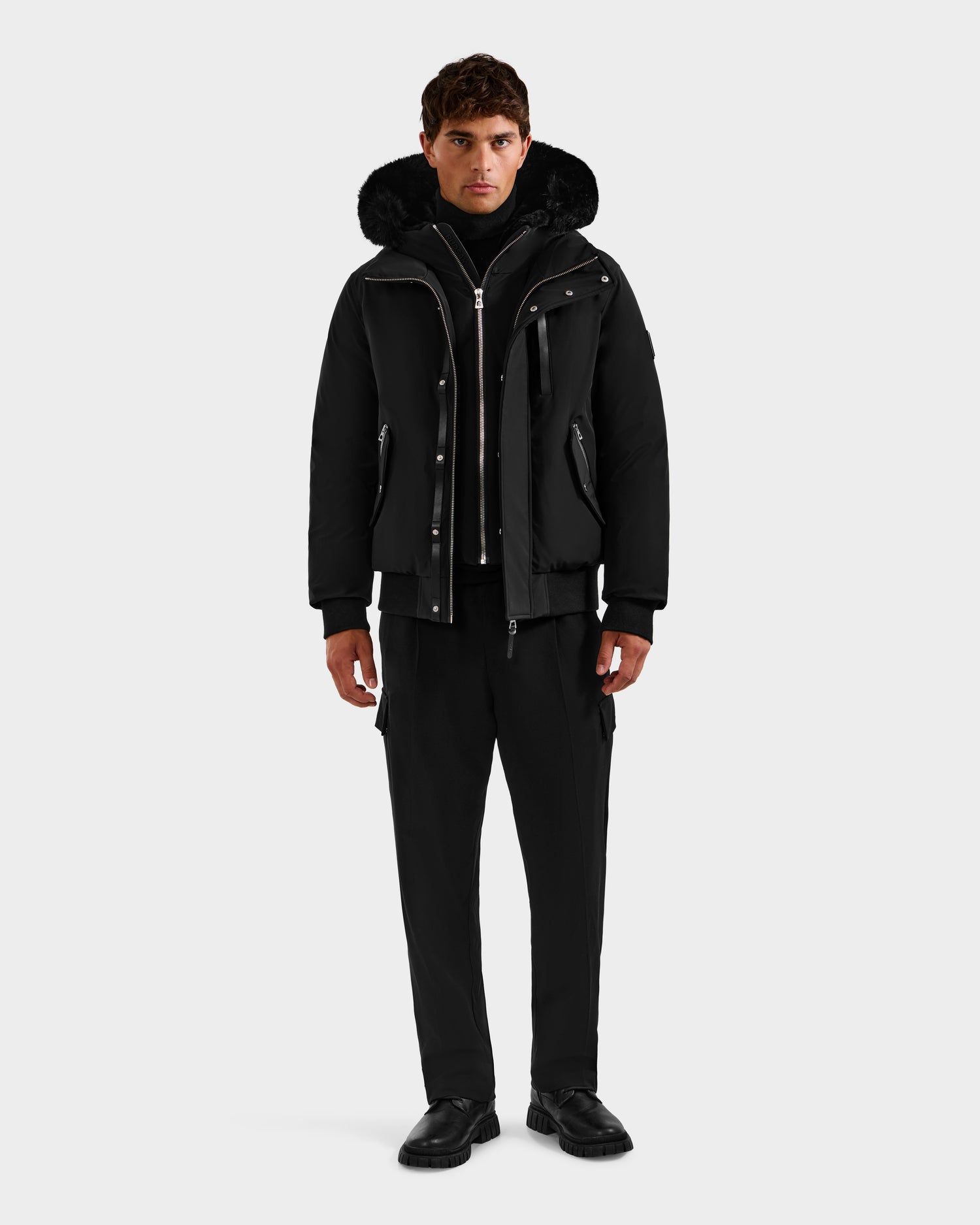 A man faces forward in a Rudsak ROCKY - 6125682 Black men's down bomber winter jacket with fur trim made from recycled nylon, paired with black pants and black shoes, against a plain white background.
