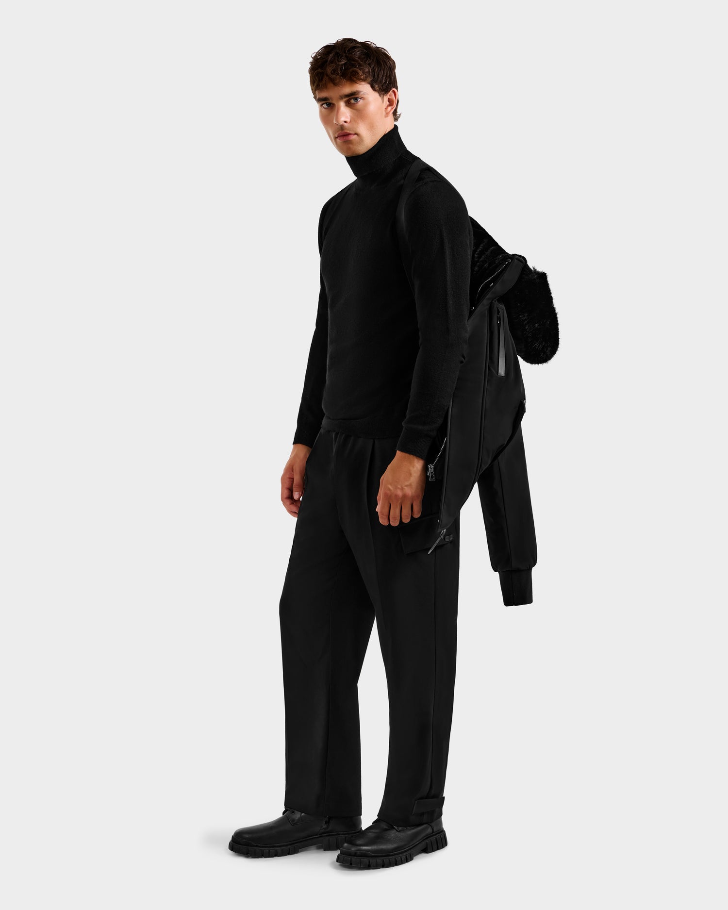 A young man stands facing slightly left, wearing black attire and holding the Rudsak ROCKY - 6125682 Black recycled nylon men's down bomber over his shoulder against a plain light gray background.