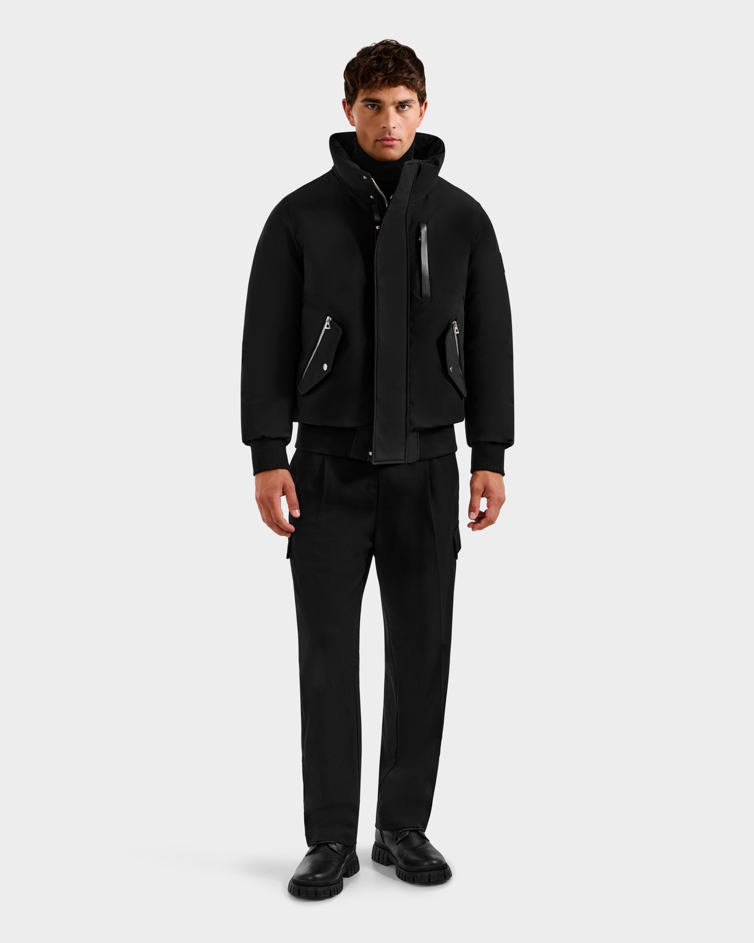 A man faces forward against a light gray background, wearing the Rudsak ROCKY - 6125682 Black men’s down bomber with zipper details, paired with black pants and shoes.