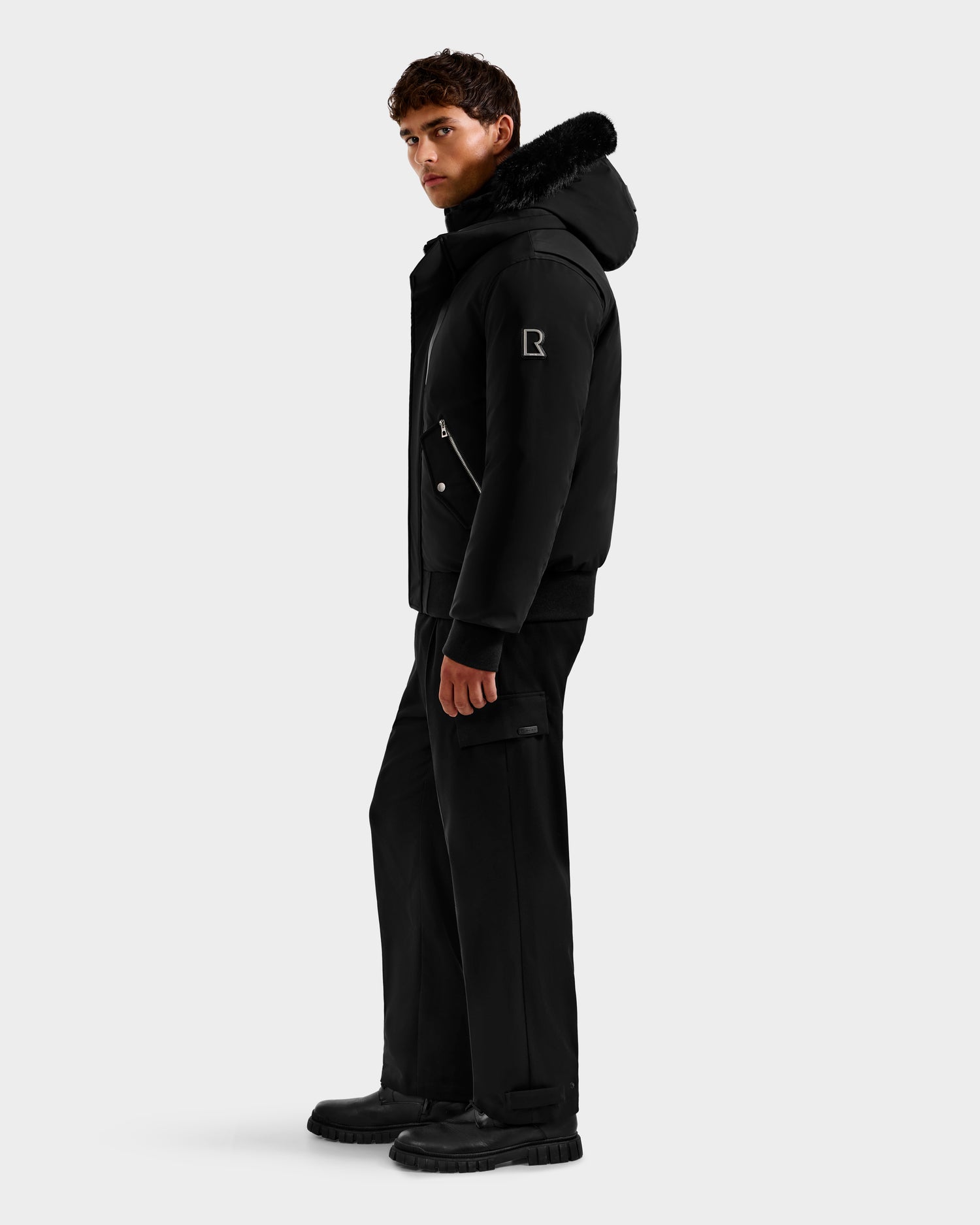 A man stands sideways against a plain light background wearing the Rudsak ROCKY - 6125682 Black recycled nylon winter bomber jacket with fur trim, paired with matching black pants and boots.
