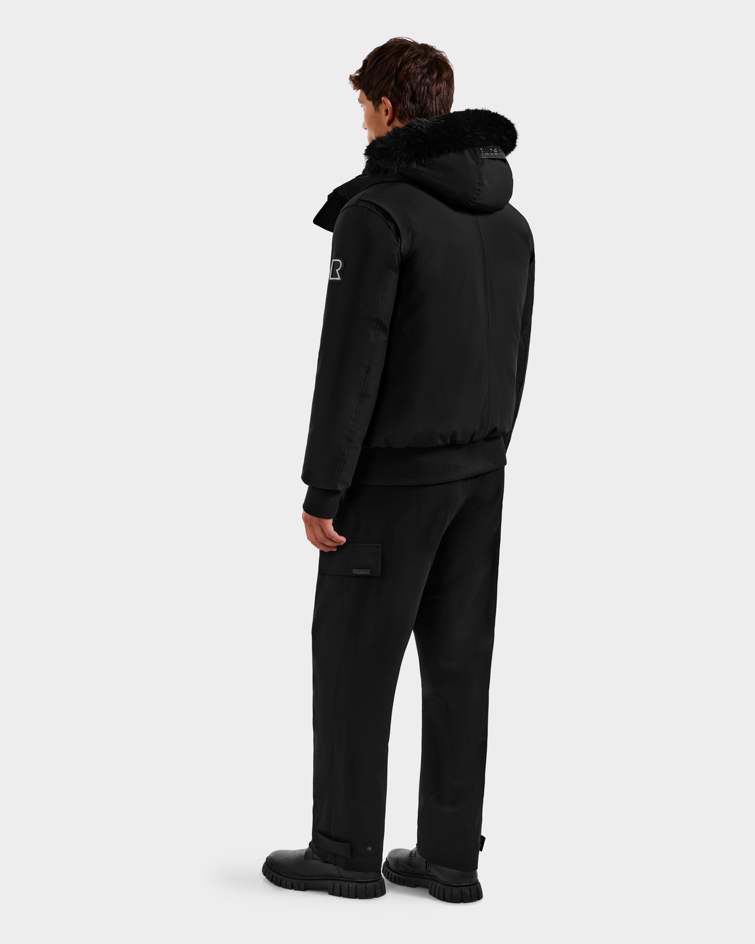 A man, seen from behind, wears the Rudsak ROCKY - 6125682 Black bomber with faux fur trim, made from recycled nylon, paired with black pants and shoes, set against a plain light gray background.