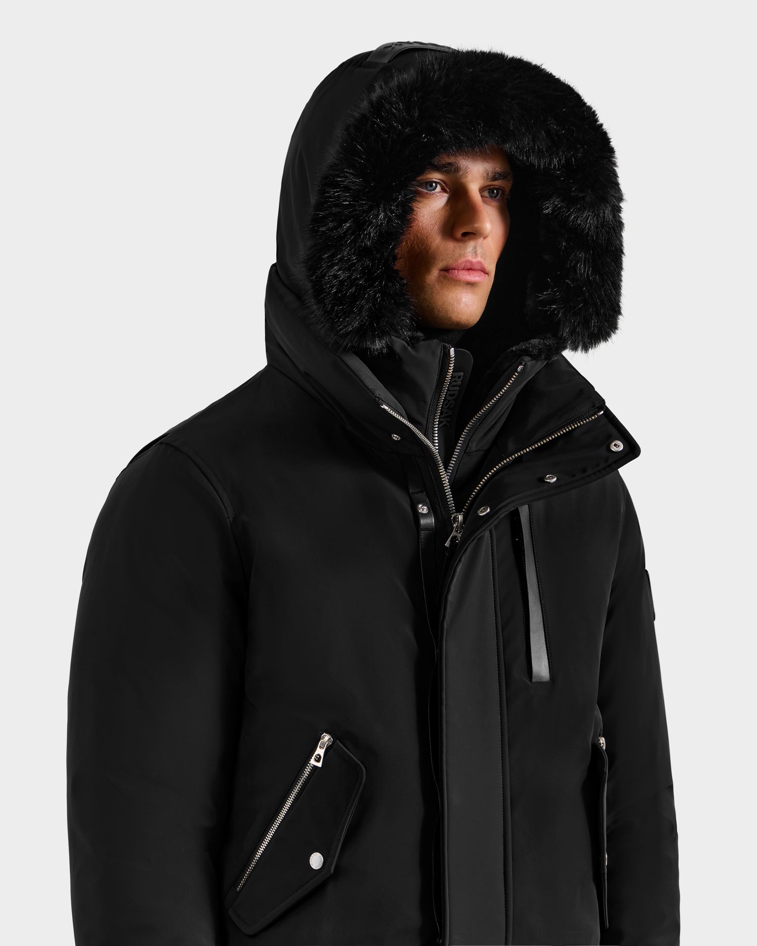 A man wears the Rudsak ROCKY - 6125682 Black, a men's winter bomber with a black faux fur-lined hood, zippered pockets, high collar, and recycled nylon construction, looking slightly to the side.