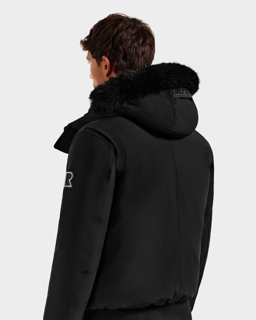 A person with short brown hair is seen from behind, wearing the Rudsak ROCKY - 6125682 Black recycled nylon winter bomber jacket with a fur-trimmed hood and sleeve patch. The background is plain light gray.