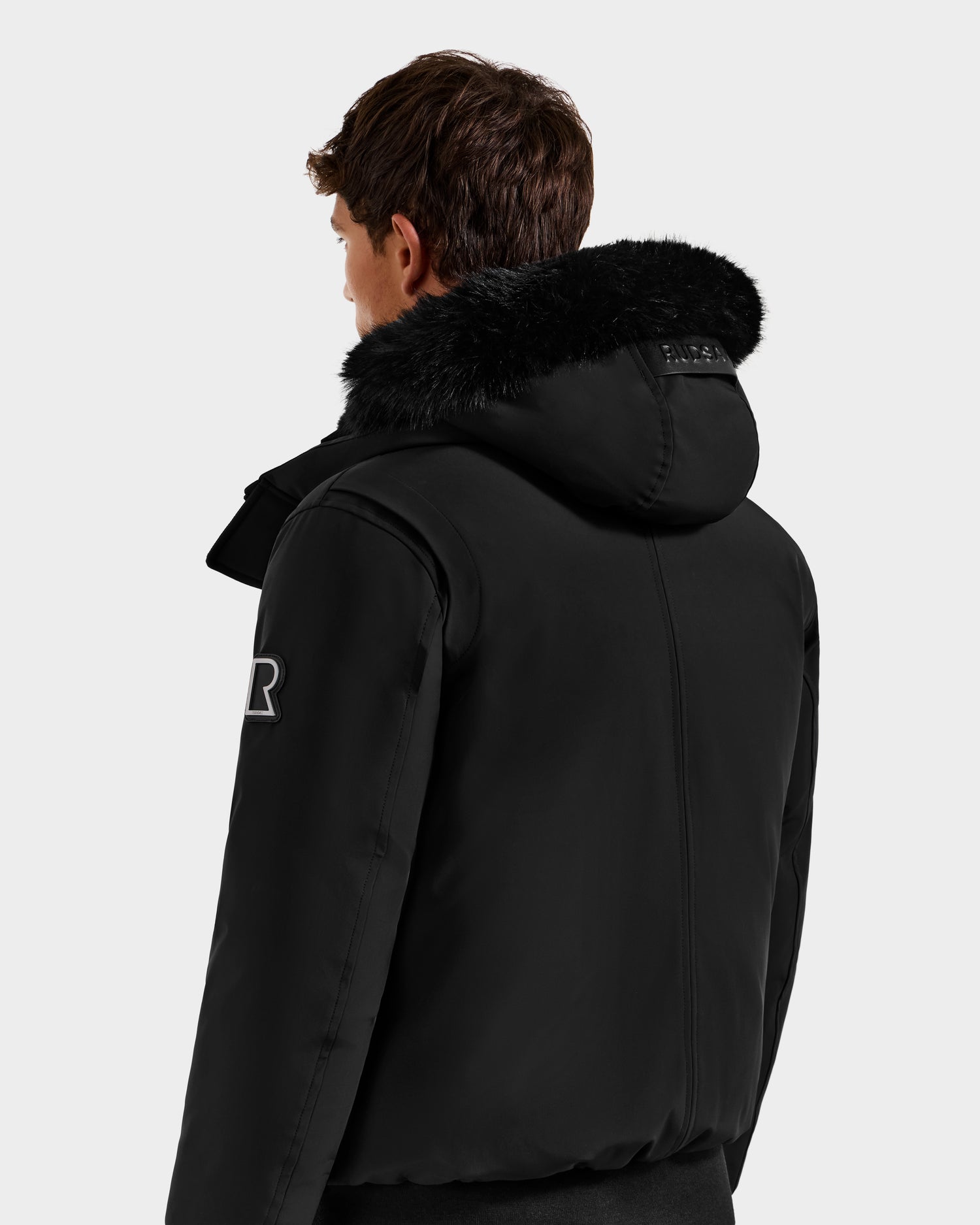 A person with short brown hair is seen from behind, wearing the Rudsak ROCKY - 6125682 Black recycled nylon winter bomber jacket with a fur-trimmed hood and sleeve patch. The background is plain light gray.