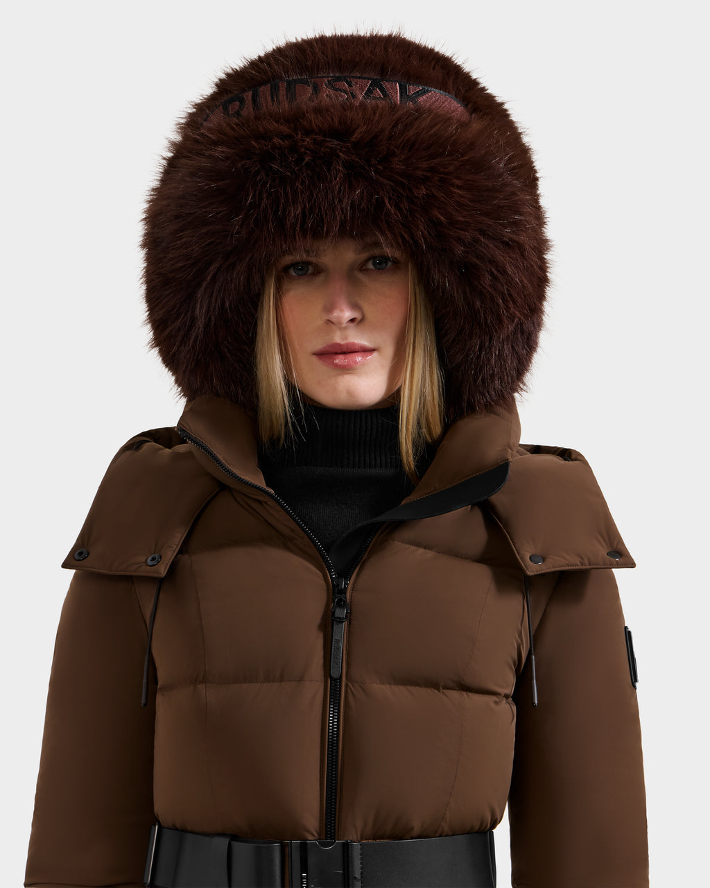 A woman with straight blonde hair and a neutral expression models the Rudsak ROMINA - 8425545 Espresso puffer jacket, featuring a large, fluffy fur hood, against a plain light background—showcasing cozy winter style.