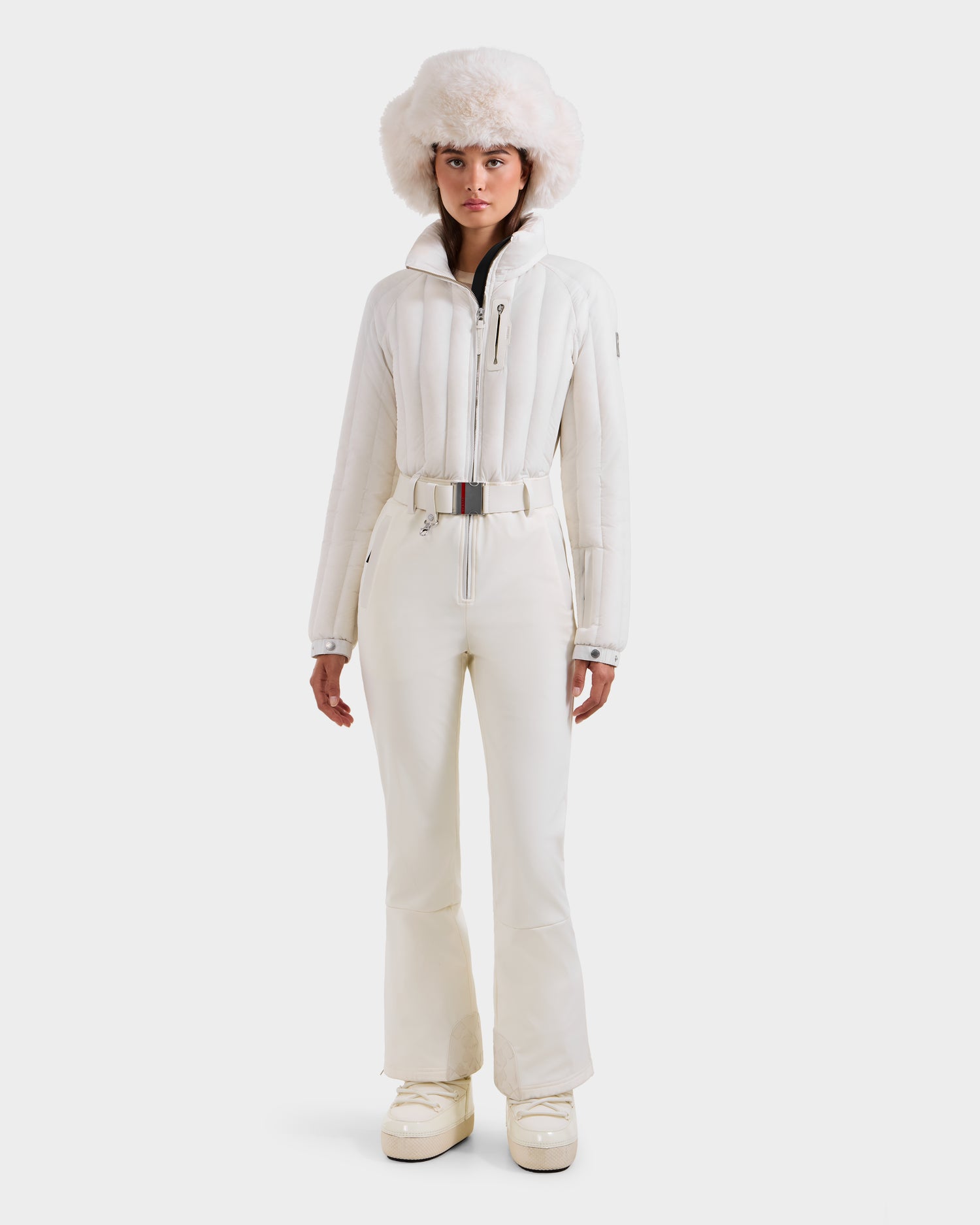 A woman faces forward in the Rudsak SAGE - 8124782 Iceland all-white ski suit with a puffy jacket, high-waisted pants, snow boots, wide belt, and large fluffy fur hat against a plain white background.