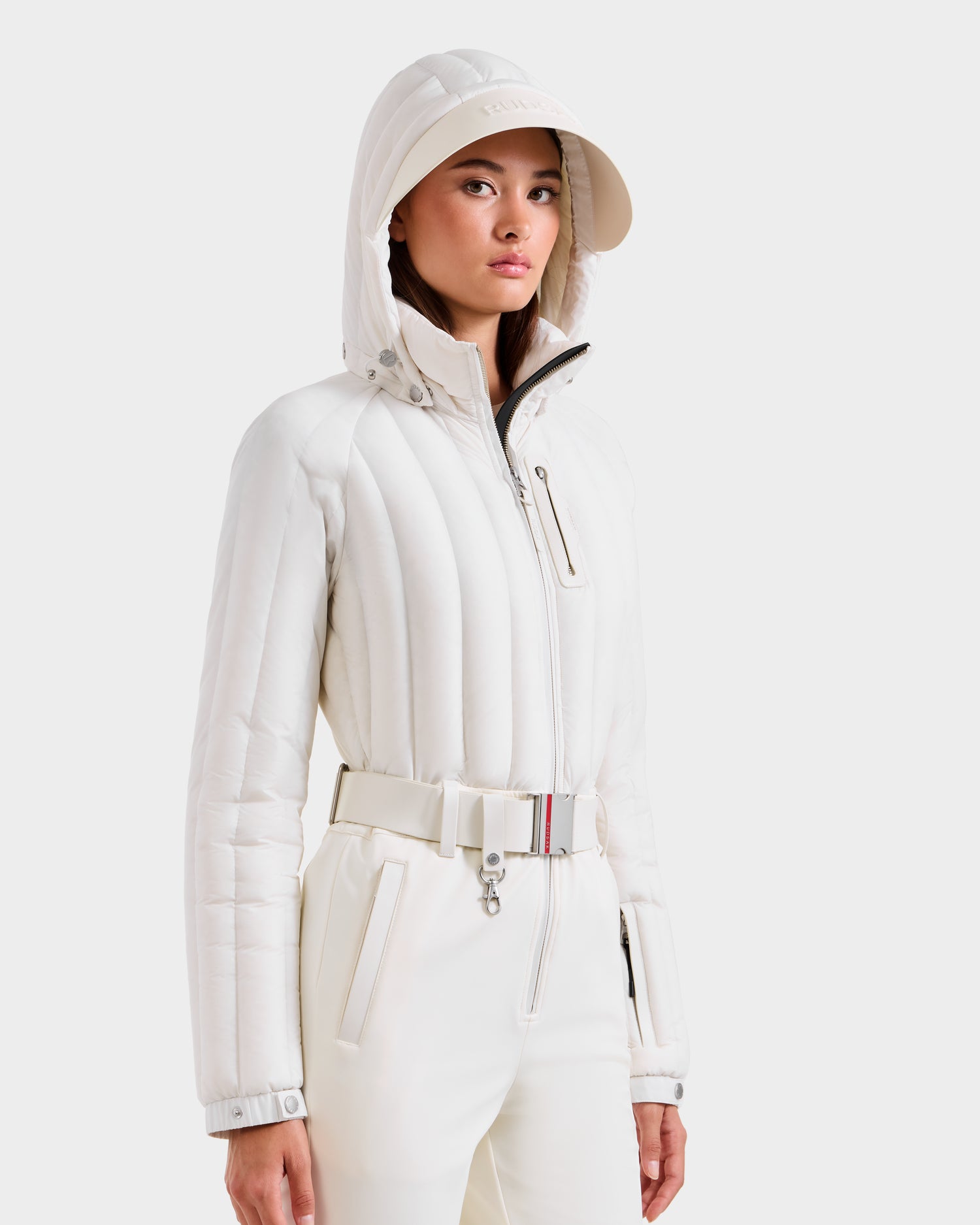 A woman wears the Rudsak SAGE - 8124782 Iceland women's ski suit in white with a hood and matching cap. The sporty, stylish outfit features quilted detailing, a zippered chest pocket, and RECCO rescue technology.