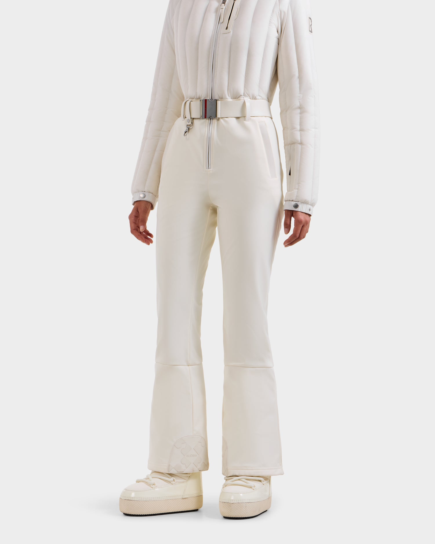 A woman stands upright against a plain white background, wearing the stylish SAGE - 8124782 Iceland one-piece ski suit by Rudsak, featuring advanced RECCO rescue technology and paired with insulated white ski boots and a matching belt.