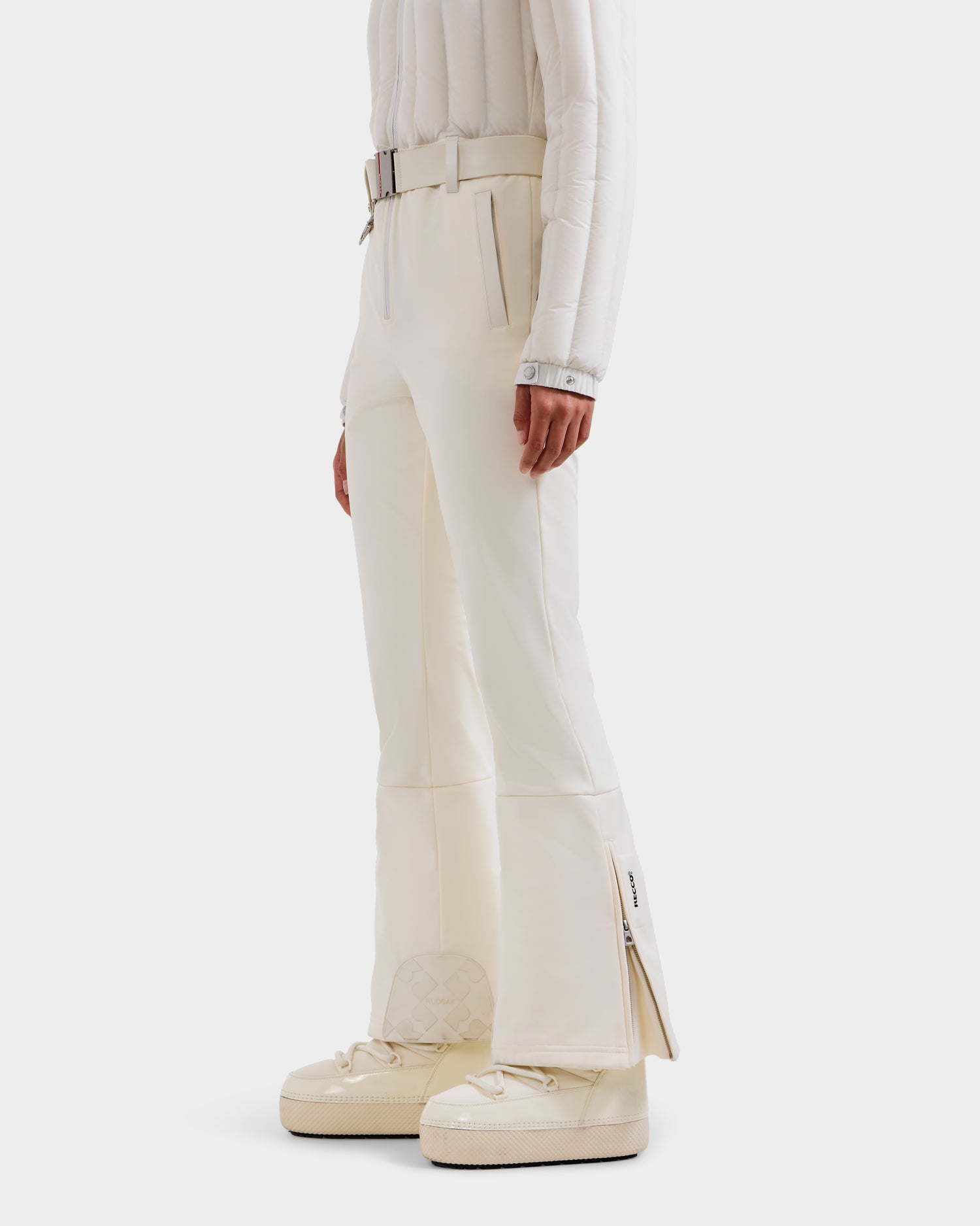 A person wears the Rudsak SAGE - 8124782 Iceland set: white flared pants with a side zipper, a belted jacket, and quilted sneakers—delivering a modern, crisp women’s ski suit vibe against a plain backdrop.