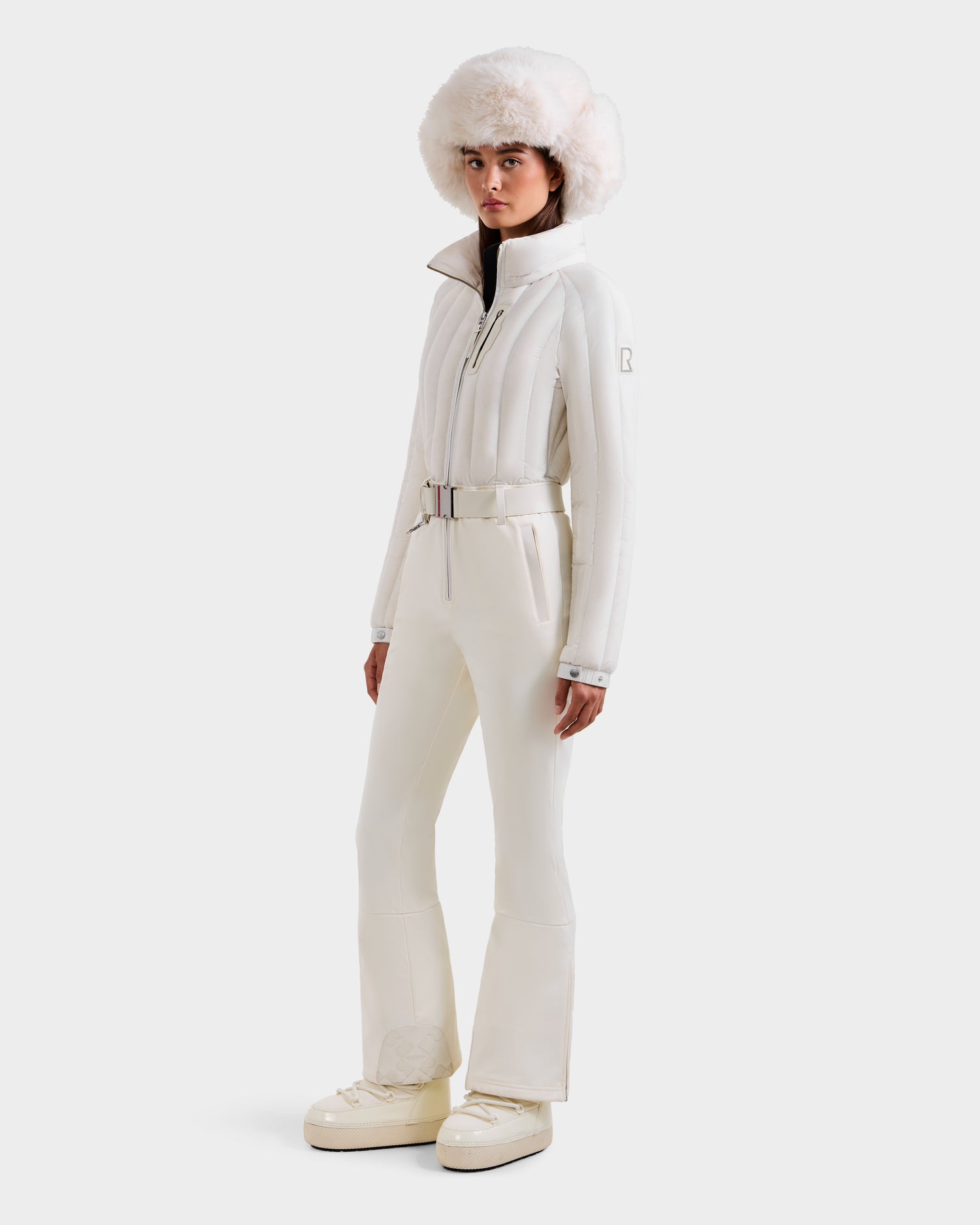 A woman models the Rudsak SAGE - 8124782 Iceland ski suit, complete with a belted jacket, flared pants, chunky snow boots, and a white fur hat—stylish and slope-ready against a plain white backdrop.